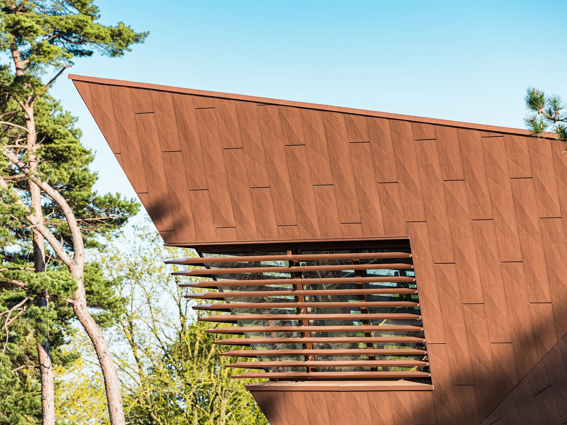 La station de pompage d'eau potable de Lottum (Pays-Bas) s'intègre harmonieusement à son environnement naturel, telle une sculpture architecturale. La façade en aluminium PREFA Siding.X, d'une teinte rouille chaleureuse, contraste de façon saisissante avec la verdure des prairies et des collines environnantes, et séduit par sa surface finement texturée et vibrante. D'élégantes ouvertures verticales apportent une touche de précision. Durables, légers et entièrement recyclables, les panneaux en aluminium PREFA impressionnent par leur haute résistance aux intempéries, leur inaltérabilité des couleurs et leur capacité à constituer une enveloppe de bâtiment durable et nécessitant peu d'entretien. Cette station de pompage d'eau potable constitue ainsi un fleuron architectural dans le domaine des infrastructures.
