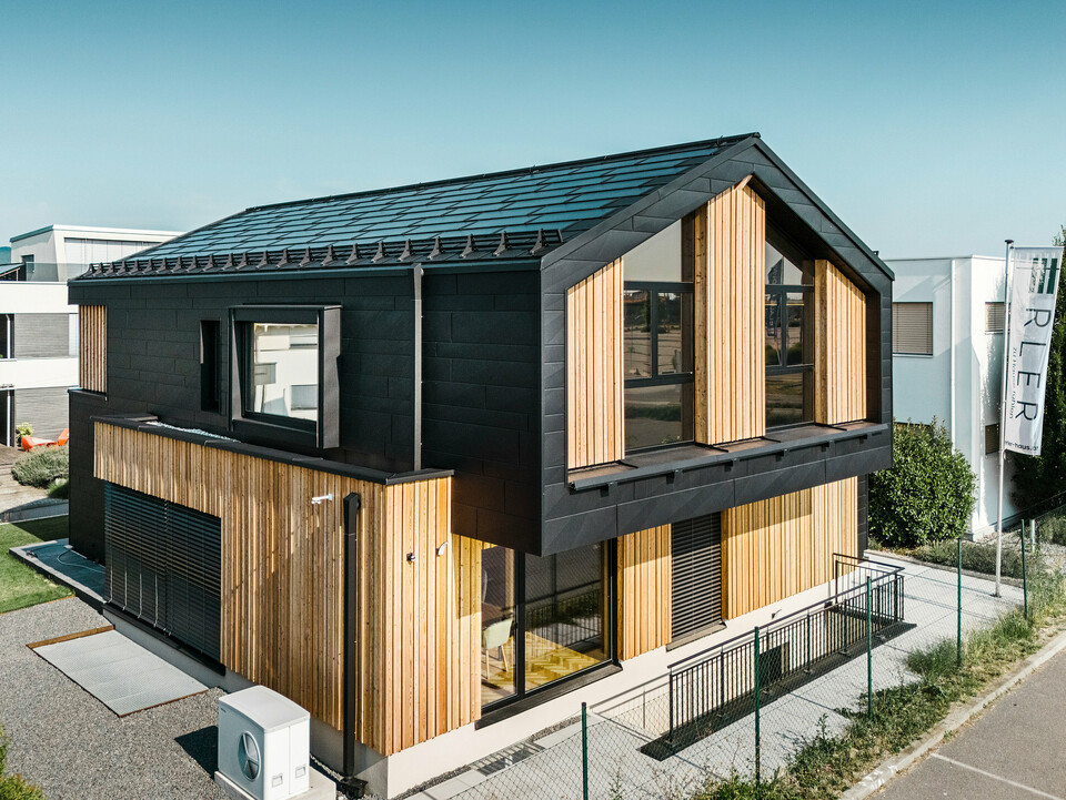 Façade de la maison modèle Autark dans le centre de maisons préfabriquées Blaue Lagune, Vösendorf (AT). Le toit est recouvert de tuiles solaires PREFA noirs et de R.16 avec un revêtement P.10 de haute qualité. La façade est constituée d'une combinaison d'éléments verticaux en bois et de PREFA Siding.X en P.10 noir, qui confère au bâtiment un caractère moderne et durable. Le drapeau de la société Erler Bau GmbH flotte sur le bord droit de l'image.
