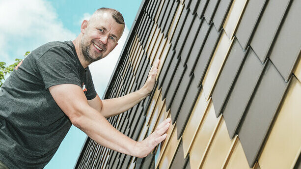 PREFA Handwerker mit grauem T-Shirt an einer PREFA Rauten-Fassade in Braun und Mayagold.