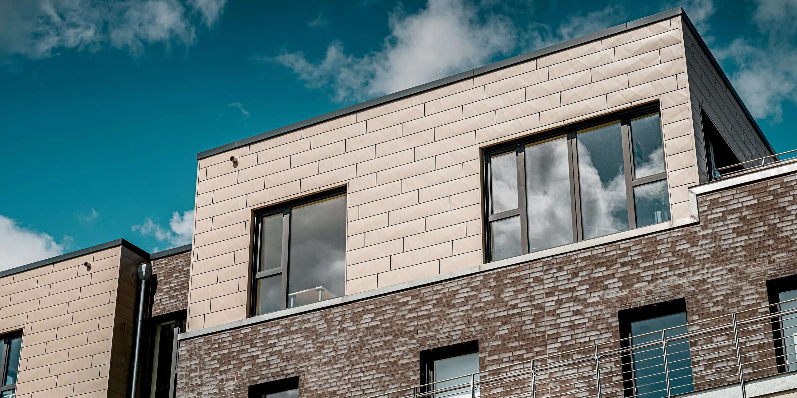 Le complexe résidentiel moderne « Hafen zur Freiheit » (Port de la Liberté) met en valeur son élégant penthouse, revêtu de panneaux Siding.X PREFA dans une chaleureuse teinte bronze. La structure caractéristique en relief des panneaux d'aluminium crée un contraste saisissant avec la façade en briques sombres. De larges fenêtres reflètent le ciel et soulignent le caractère lumineux et haut de gamme du bâtiment. Robustes et résistants aux intempéries, les éléments de façade PREFA sont particulièrement durables en bord de mer et confèrent au complexe son allure contemporaine et distinctive.