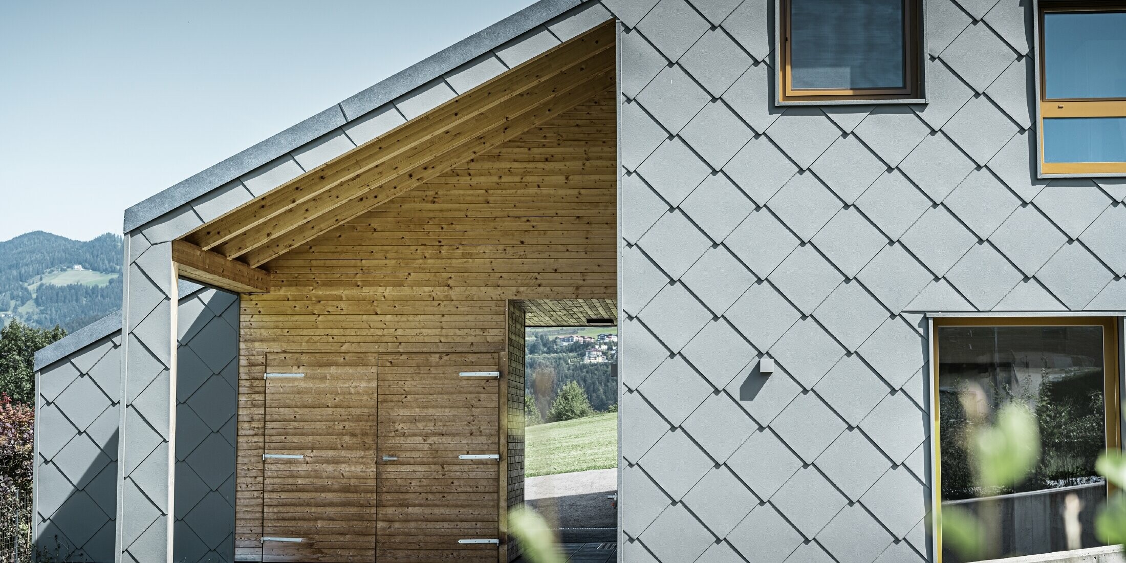 Der überdachte Eingangsbereich ist mit waagrechter Holzverkleidung gemacht, der Rest der Fassade ist mit der großen PREFA Wandraute aus Aluminium in Hellgrau verkleidet