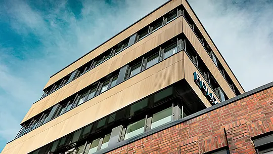 Vue dynamique du dessous du commissariat 43 à Hambourg : ce bâtiment moderne aux lignes épurées impressionne par sa façade en aluminium PREFA, composée de profils extrudés « triangle » d’un élégant bronze moyen. La surface finement nervurée attire le regard et crée de captivants jeux d’ombre et de lumière le long des bandes horizontales des fenêtres. Contrastant avec la brique massive, elle affirme une forte identité architecturale. L’aluminium PREFA se distingue par sa résistance à la corrosion, sa durabilité et son faible besoin d’entretien – un matériau idéal pour une conception de façade durable et précise.