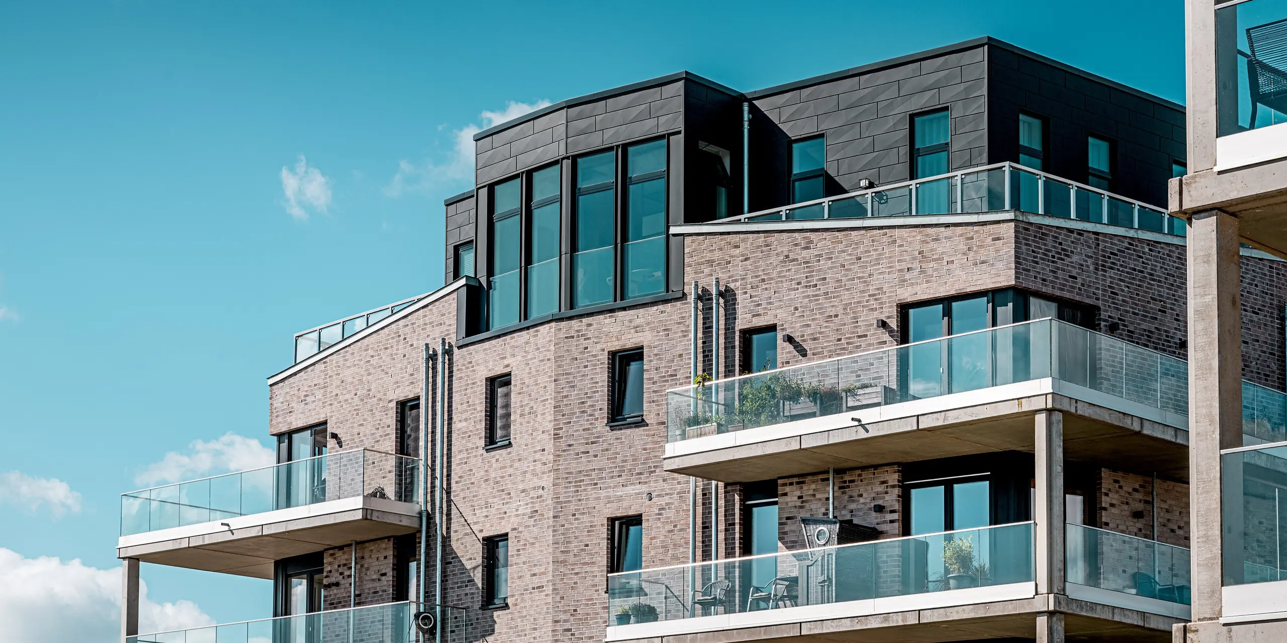 Le complexe résidentiel moderne « Hafen zur Freiheit » (Port de la Liberté) présente une façade remarquable, visible dans la partie supérieure de l'image. Elle associe des briques de parement claires à l'élégant revêtement PREFA Siding.X couleur P.10 Anthracite. Les surfaces en aluminium à la structure audacieuse qui entourent le dernier étage attirent particulièrement le regard et confèrent au bâtiment une allure dynamique, presque sculpturale. Les généreux balcons vitrés reflètent la lumière et soulignent la qualité architecturale. De plus, les panneaux en aluminium PREFA, résistants aux intempéries, offrent une protection optimale malgré l'exposition du littoral et valorisent le bâtiment sur le long terme.