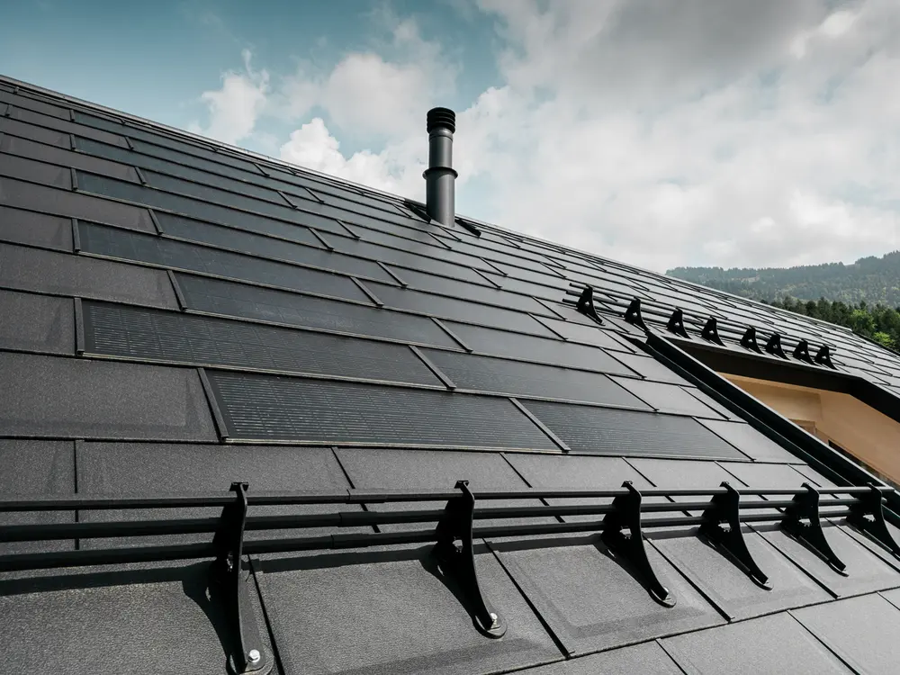 Gros plan sur le toit d'une maison moderne, recouvert de panneaux solaires noirs PREFA et de panneaux de toiture R.16. Des grilles pare-neige soulignent la fonctionnalité du toit. Une fenêtre de toit et une cheminée d'évacuation rompent l'uniformité de la surface du toit, qui s'étend sur un fond de collines boisées et un ciel bleu clair avec quelques nuages.