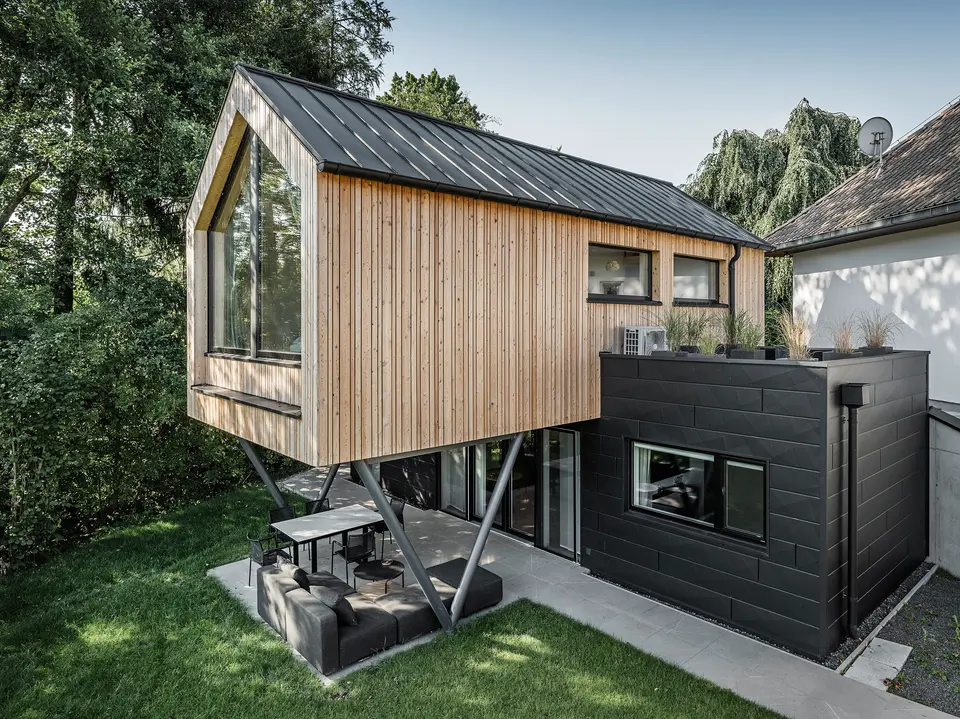 Das 'Woodstock Chalet 5' beeindruckt mit einer eindrucksvollen Kombination aus natürlichen Holzoberflächen und den modernen PREFA Siding.X Fassadenelementen in Anthrazit. Die überhängende Holzstruktur des Obergeschosses ragt markant über die dunklen Fassadenelemente, die das Erdgeschoss verkleiden, und erzeugt so ein Spiel aus Licht und Schatten. Die klaren Linien und die robuste Konstruktion der Siding.X Elemente ergänzen das innovative Design des Gebäudes und bieten einen spannenden visuellen Kontrast zur umgebenden Vegetation und dem traditionellen Gebäude im Hintergrund. Am Dach des Chalets wurde das PREFALZ Dachsystem in Form einer gleichmäßigen Stehfalzdeckung verlegt. Am rechten Rand des Erdgeschosses ist das PREFA Dachentwässerungssystem zu erkennen, das analog zur Aluminiumfassade in Anthrazit gehalten ist. Für die Dachentwässerung wurden der Wasserfangkasten gefolgt vom Ablaufrohr montiert.