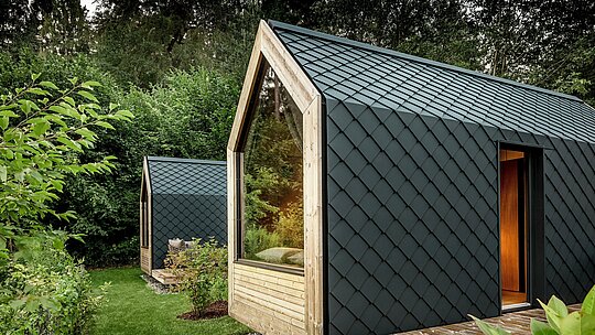 Deux maisons minimalistes de Secret Koi Garden se nichent dans la végétation luxuriante au bord du lac Natterer. Leurs façades et toitures sont entièrement revêtues de petits losanges en aluminium PREFA P.10 anthracite, créant une texture saisissante évoquant des écailles de poisson. De larges surfaces vitrées et des détails en bois chaleureux contrastent harmonieusement avec l'aluminium sombre. Durable et résistant aux intempéries, l'aluminium PREFA assure une protection fiable et souligne le caractère haut de gamme de cette architecture de glamping.