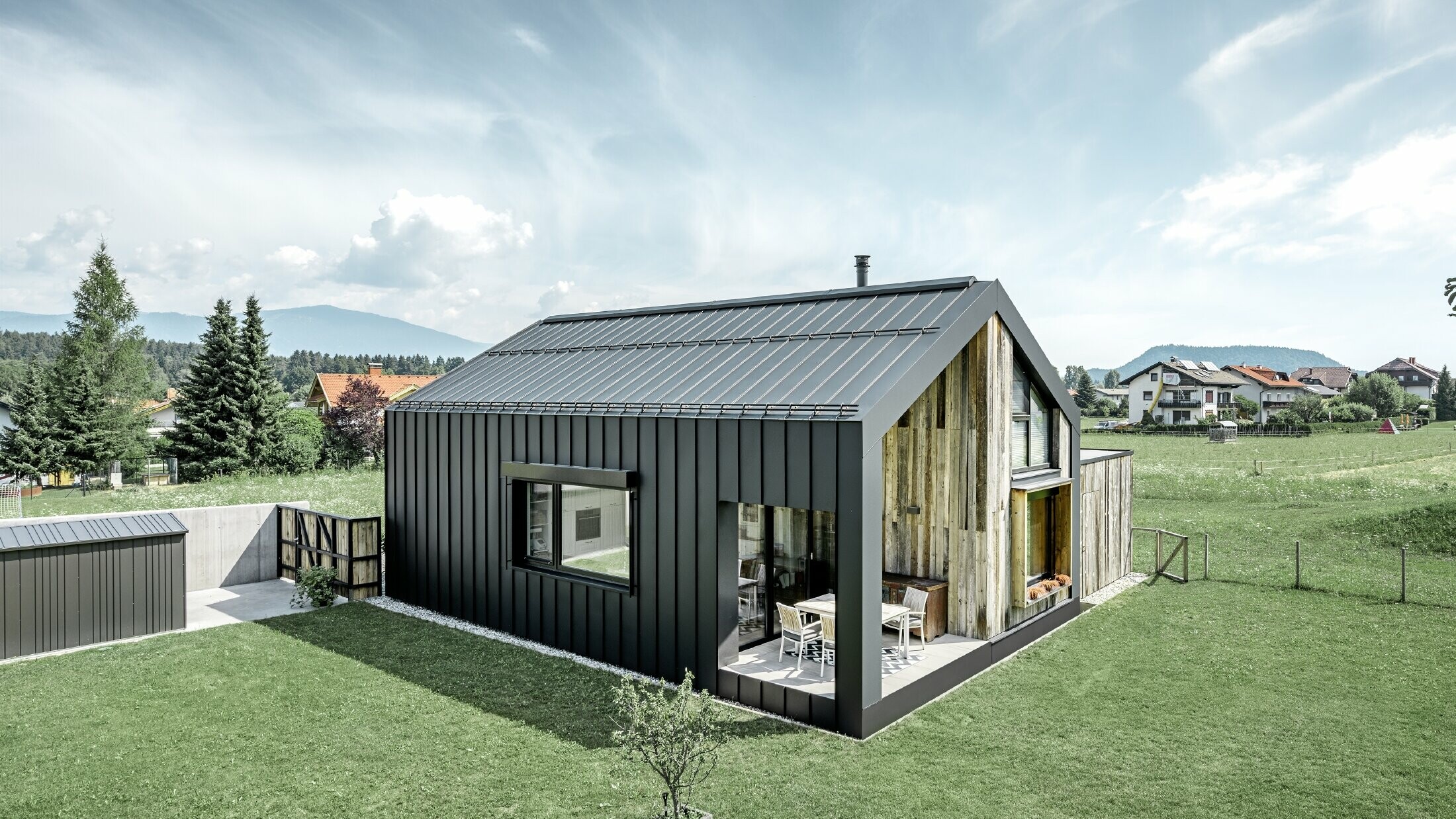 Maison individuelle avec toit à pignon ; la toiture et la façade sont recouvertes avec PREFALZ de PREFA en P.10 anthracite. Les bacs de la surface de toit se prolongent jusque sur la façade. Une partie de la façade est revêtue de bois patiné.