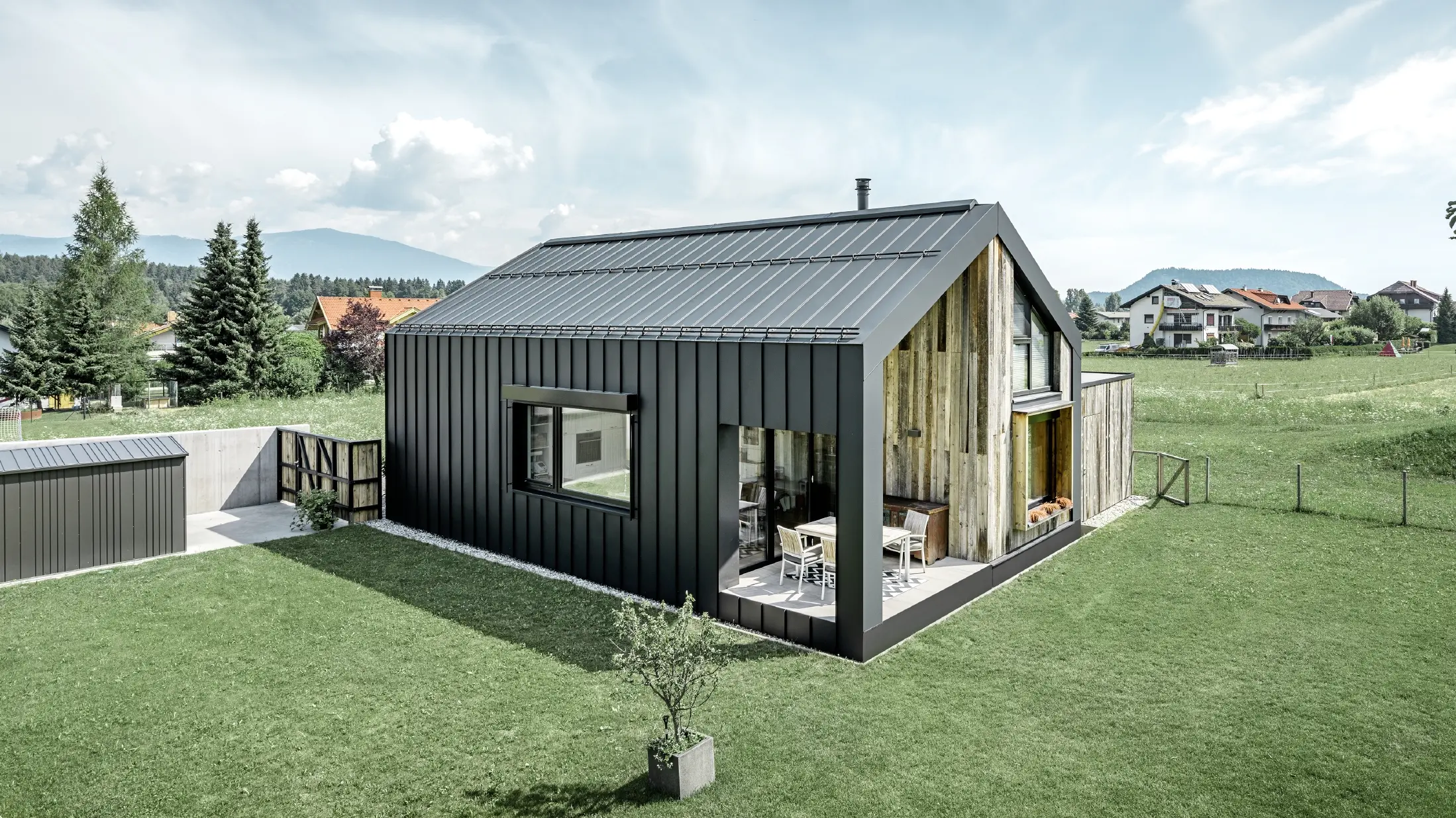 Maison individuelle avec toit à pignon ; la toiture et la façade sont recouvertes avec PREFALZ de PREFA en P.10 anthracite. Les bacs de la surface de toit se prolongent jusque sur la façade. Une partie de la façade est revêtue de bois patiné.