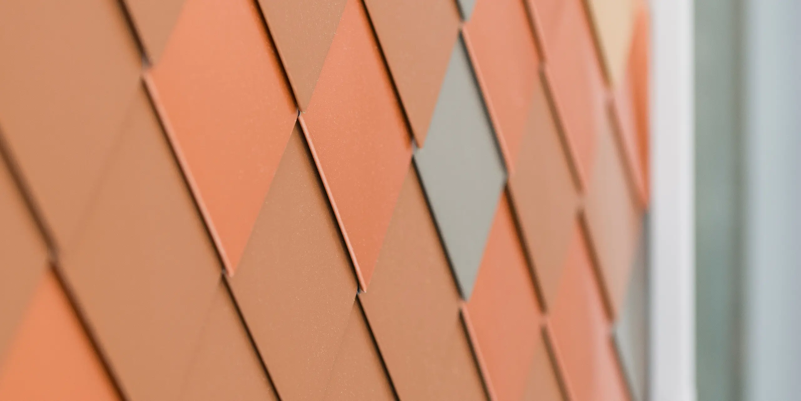Vue détaillée d'une façade moderne en aluminium PREFA avec un motif géométrique de différentes couleurs. Les petites pièces en aluminium en FALZONAL sont disposées en forme de losange et créent un aspect tridimensionnel qui met en valeur la structure et la profondeur des couleurs.
