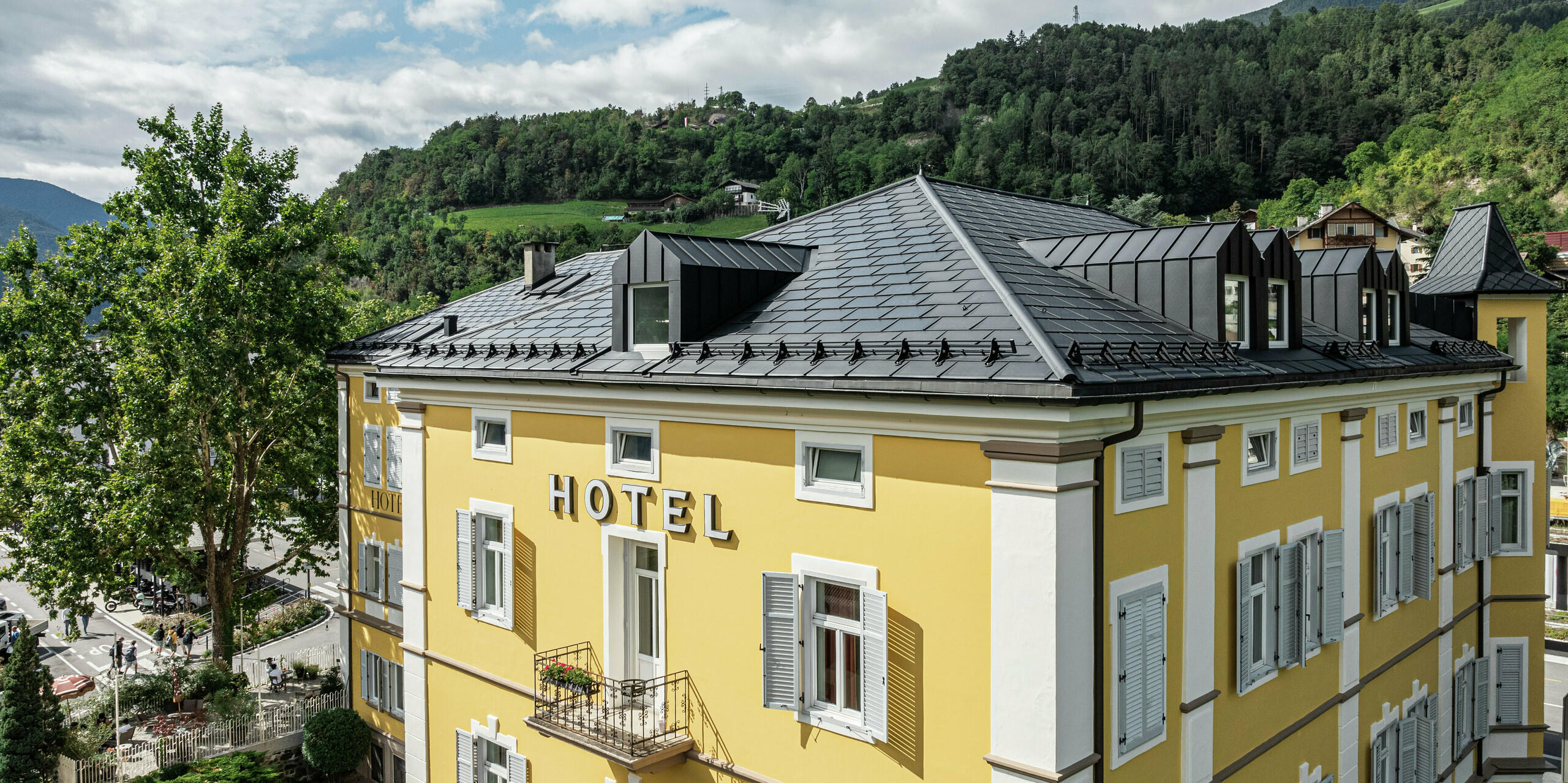 Prise de vue par drone de l'hôtel Jarolim à Brixen avec un toit en aluminium durable composé de tuiles solaires PREFA et de R.16 dans un look noir élégant avec un revêtement P.10 de haute qualité. Le toit PREFA robuste et résistant aux intempéries allie esthétique et fonctionnalité et est idéal pour les bâtiments classés. L'intégration harmonieuse des modules photovoltaïques dans les panneaux de toiture robustes en aluminium permet une production d'énergie durable sans compromettre l'architecture historique. La finition précise souligne le caractère de haute qualité des produits PREFA.