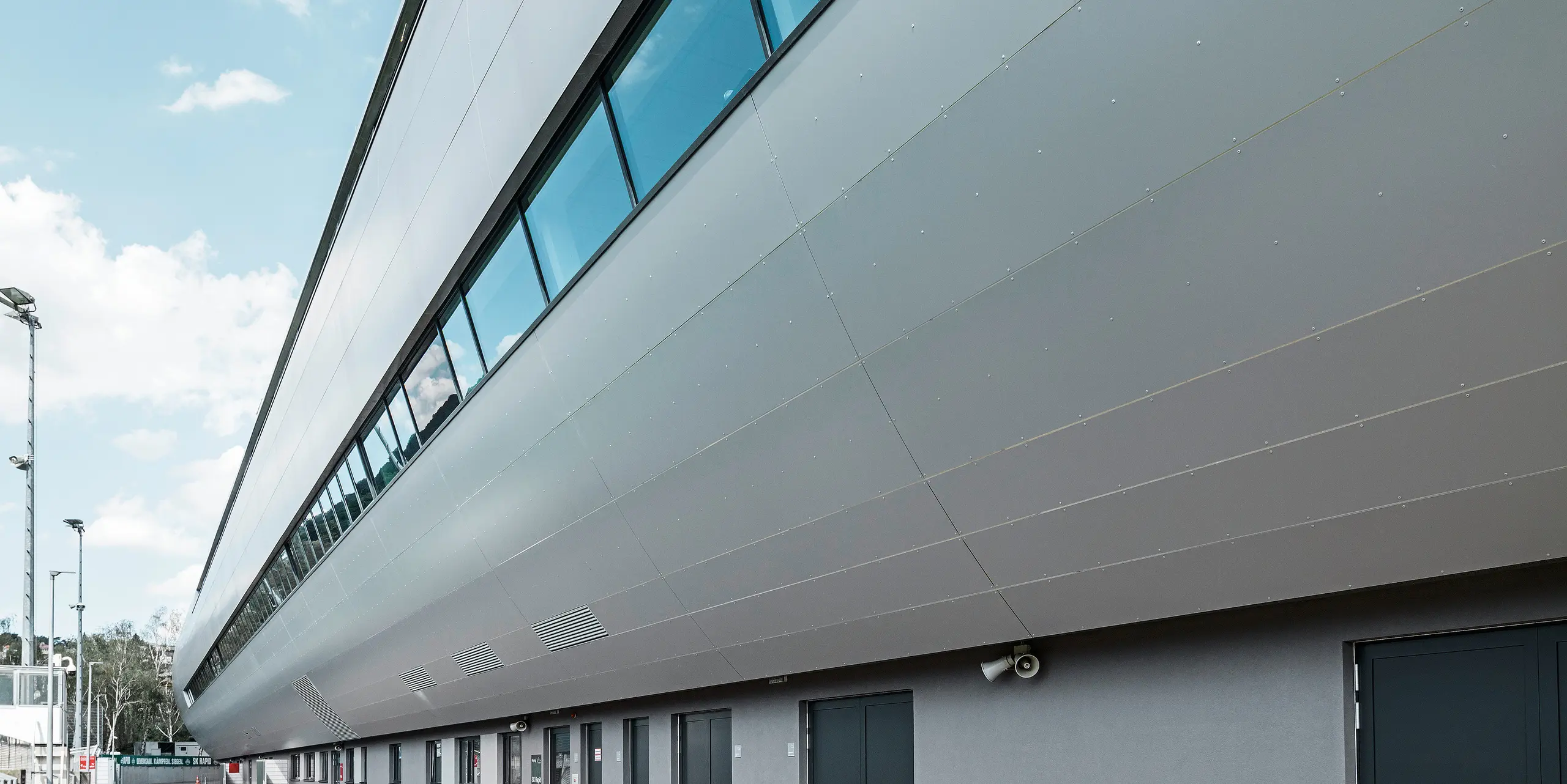 Seitenansicht des Allianz Stadions von SK Rapid Wien, verkleidet mit PREFA Aluminium Verbundplatten in Silbermetallic. Die Fassade zeigt die beeindruckende Länge und klare Linienführung des Bauwerks, während die großflächigen Fenster den Blick zum Trainingsplatz rahmen. In den Fensterscheiben reflektiert der blaue Himmel. Die hochwertige Metallverkleidung verkörpert Beständigkeit und moderne Architektur.