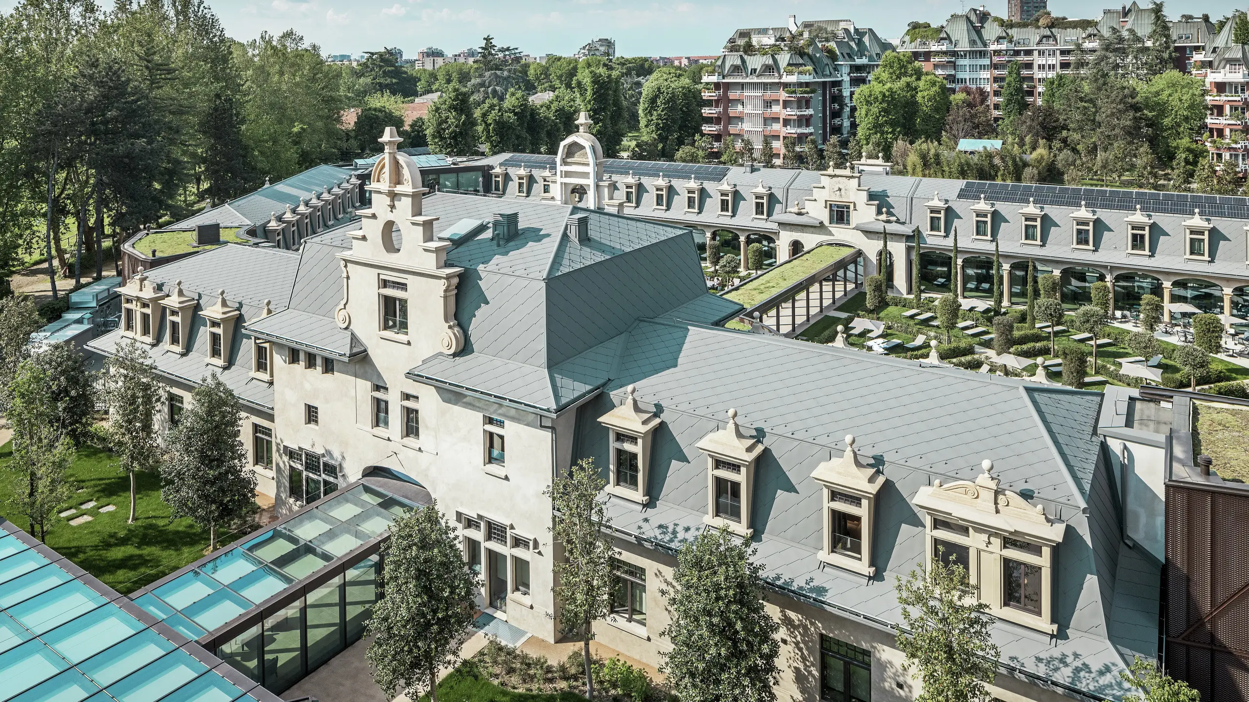 Vue d'ensemble de l'impressionnant complexe « De Montel – Terme Milano », alliant architecture Art nouveau classée et ambiance bien-être moderne. La toiture généreuse en losanges PREFA 44 × 44 P.10 gris souris confère au complexe une élégante uniformité. Les modules photovoltaïques intégrés, les toitures végétalisées et les liaisons vitrées entre les bâtiments témoignent de l'engagement en faveur du développement durable et du design contemporain en contexte urbain.