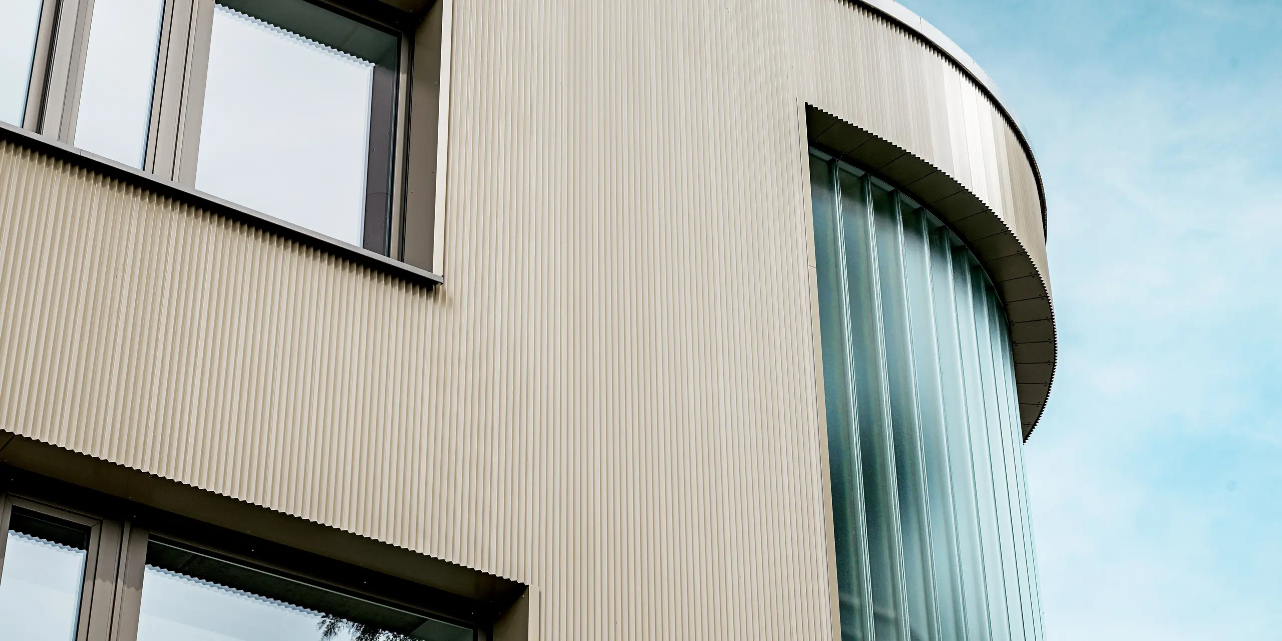 L'image révèle une section fascinante de la façade du bâtiment Festo à Langenhagen, revêtue de profils extrudés triangle PREFA. La courbe douce du bâtiment et les surfaces vitrées verticales à l'angle créent un contraste saisissant avec la façade en aluminium texturé. Les lignes épurées des profils triangle de couleur bronze et leur harmonie avec les généreuses fenêtres et les éléments vitrés confèrent au bâtiment une allure élégante et moderne. Cette combinaison de matériaux fonctionnels et de design esthétique garantit un résultat architectural durable, nécessitant peu d'entretien et d'une grande beauté visuelle.