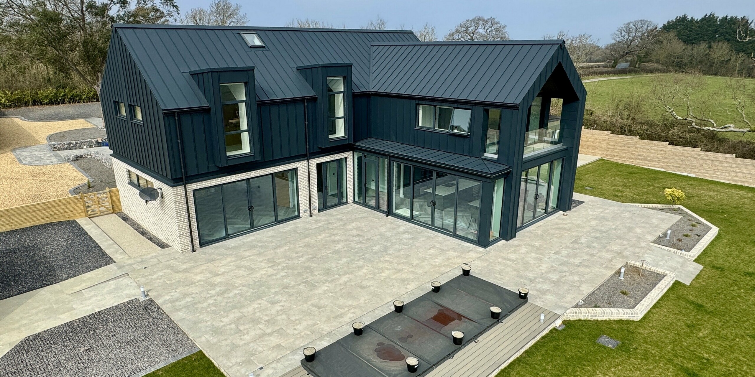 Modernes Einfamilienhaus mit markantem PREFALZ Dach und Fassade in P.10 Dunkelgrau, kombiniert mit heller Steinverkleidung im Erdgeschoss. Große Glasfronten öffnen den Wohnbereich zur weitläufigen Terrasse mit Pool. Die Aluminiumoberflächen sind langlebig, korrosionsbeständig und ideal für die Küstenlage. Das Zusammenspiel von dunklem Dach, Glas und Naturstein schafft einen eleganten Kontrast und unterstreicht die hochwertige Architektur des Projekts Bullenmead auf der Isle of Wight.