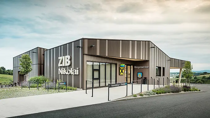 Vue de face du centre événementiel ZIB Nikolai à St. Veit, avec une façade moderne en aluminium à rayures marron-beige-gris et une grande inscription « ZIB Nikolai ». La zone d'entrée présente une signalisation claire pour les événements, le stationnement et les vélos, ainsi qu'un service citoyen intégré et une succursale de la Raiffeisenbank. En arrière-plan, des collines verdoyantes et un vaste paysage s’étendent sous un ciel nuageux.
