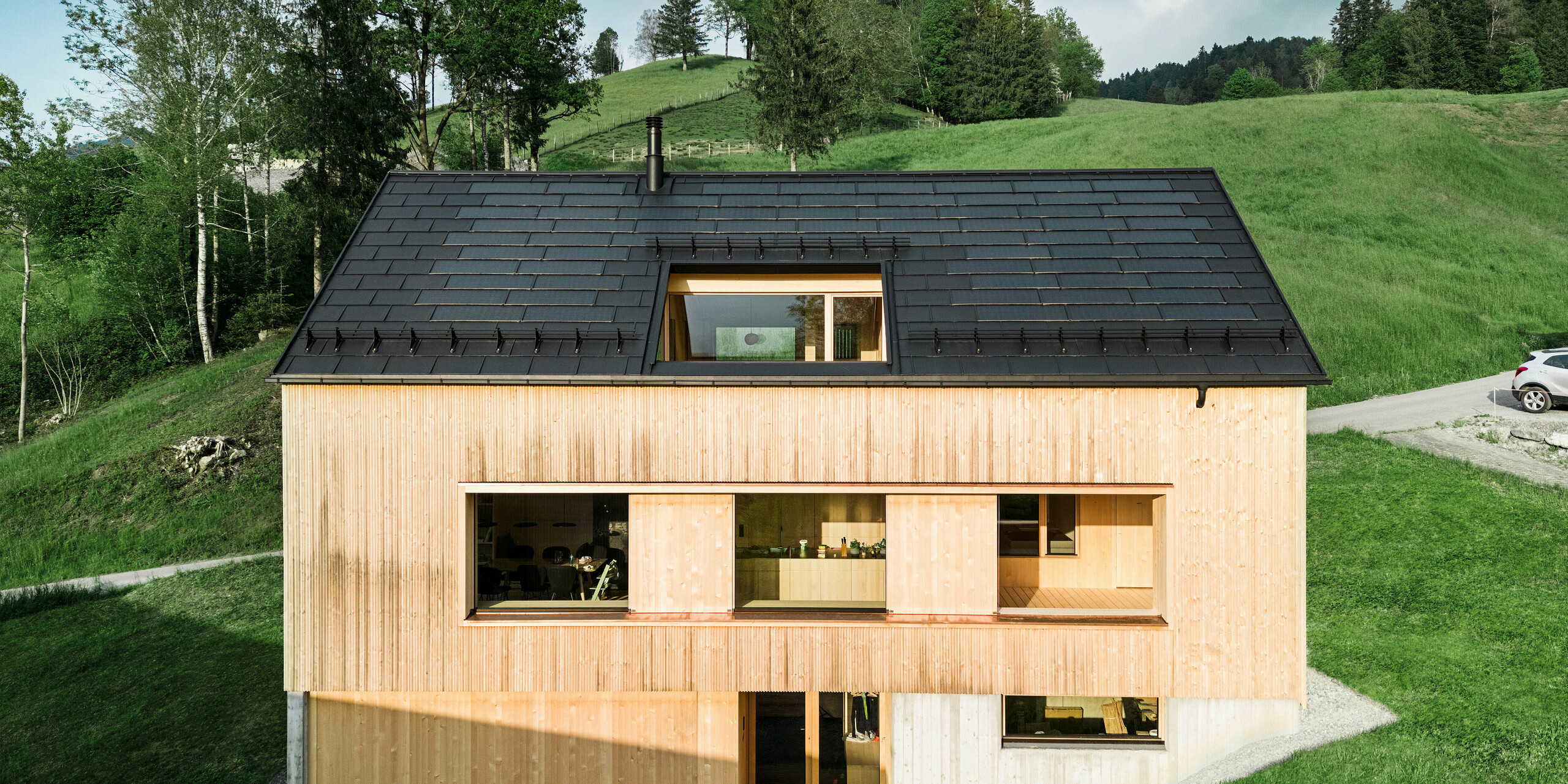 Écologique et esthétique : la photo montre une maison écologique moderne à Egg, équipée de la tuile solaire innovante PREFA. Le toit en aluminium robuste et noir profond, réalisé avec les R.16 PREFA, se fond visuellement dans l'environnement naturel et souligne les lignes claires de l'architecture contemporaine en bois. Les modules solaires intégrés s'intègrent discrètement dans le toit et démontrent une construction durable alliant efficacité énergétique et design. Les tuiles solaires innovantes PREFA créent une symbiose entre fonctionnalité et design. Ce concept de vie durable combine des tuiles en aluminium de haute qualité avec une technologie solaire avancée et s'intègre parfaitement dans le paysage montagneux idyllique tout en minimisant l'empreinte écologique de la maison.