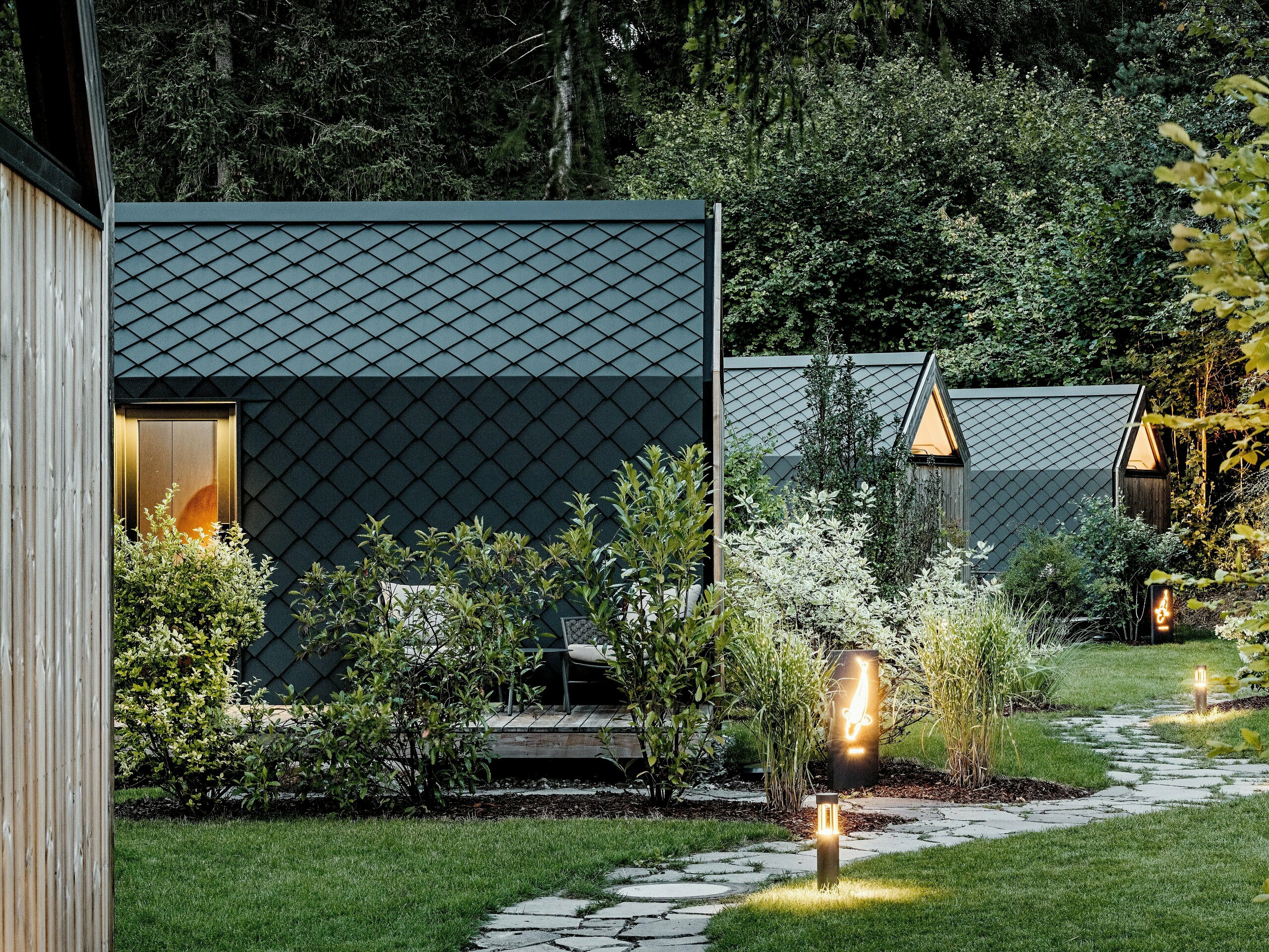 Un chemin de jardin illuminé traverse le complexe de glamping Secret Koi Garden, situé sur les rives du lac Natterer, et mène à plusieurs mini-maisons minimalistes. Ces structures compactes sont entièrement revêtues de losanges de toiture et de façade en aluminium PREFA P.10 anthracite, dont la texture en écailles de poisson crée un effet élégant sous la lumière du soir. Nichées dans une végétation luxuriante, les surfaces en aluminium PREFA, durables et résistantes aux intempéries, allient une architecture de haute qualité à la nature, à la tranquillité et à un design durable.