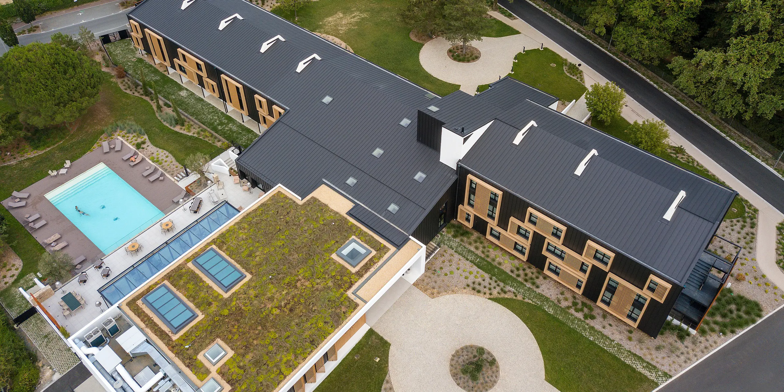 Vue aérienne de la zone d'entrée de l'Hôtel Parc du Landreau aux Herbiers, France. Le bâtiment a été revêtu du système de toiture et de façade PREFALZ en gris noir. Les cadres en bois frappants autour des fenêtres complètent le revêtement moderne en aluminium. L'espace extérieur avec piscine et toit partiellement végétalisé reflète la construction durable de l'hôtel.