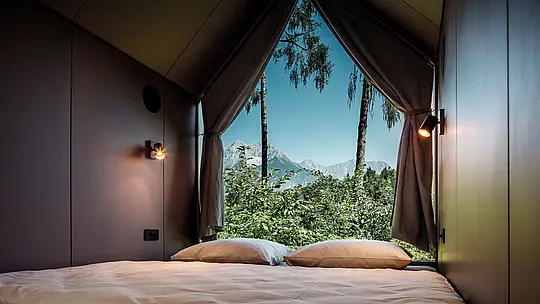 Vue depuis le lit d'une tiny house minimaliste dans le Secret Koi Garden, au bord du lac Natterer : la grande baie vitrée triangulaire ouvre la pièce sur la nature environnante, avec ses arbres et la vue sur les montagnes tyroliennes. L'intérieur épuré invite à la contemplation et souligne l'importance de ralentir le rythme. L'architecture est protégée par des losanges en aluminium PREFA, un matériau durable qui allie design, résistance aux intempéries et durabilité.