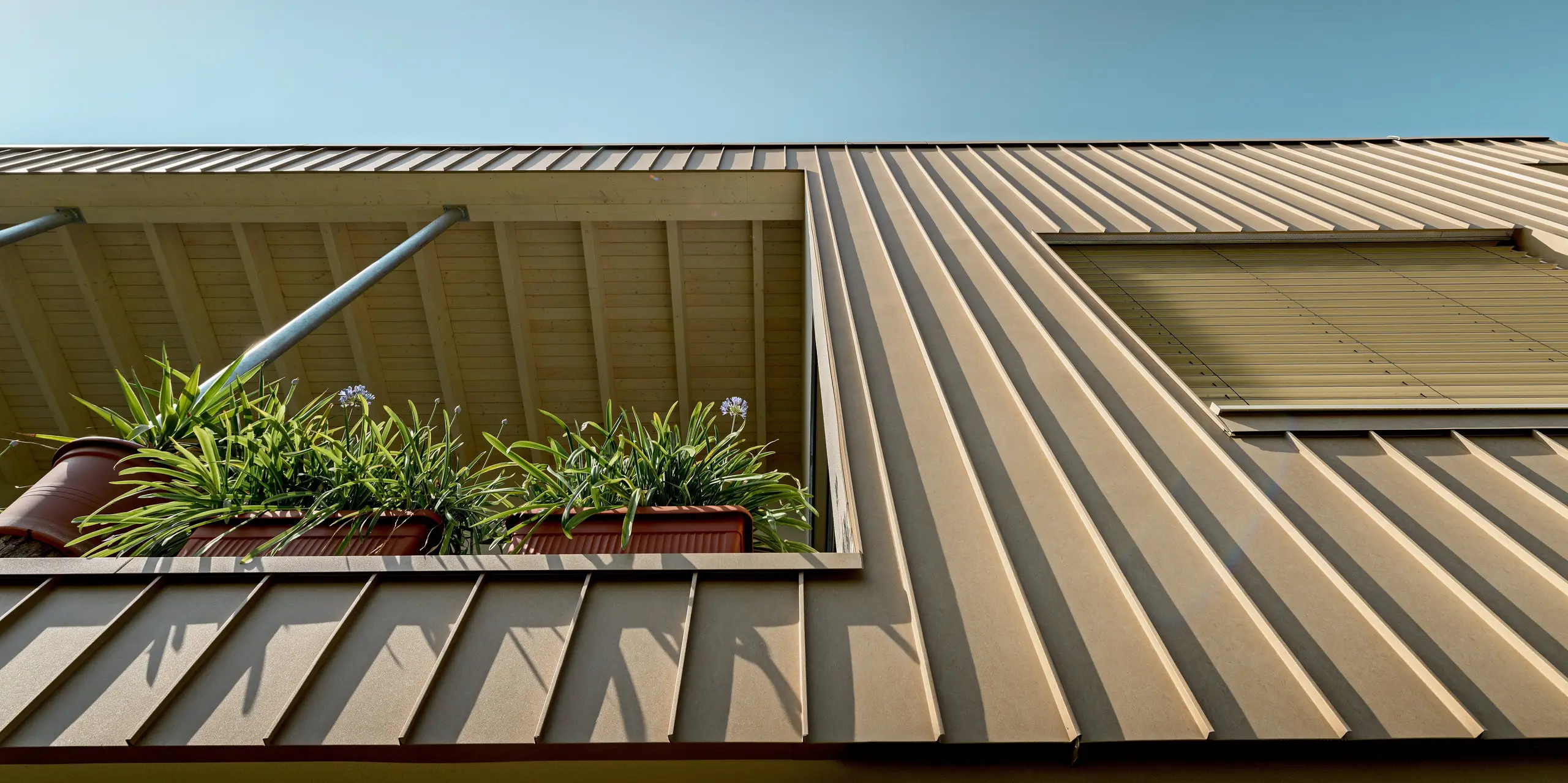 Detailaufnahme eines modernen Einfamilienhauses in Schiefling am See mit markanter PREFALZ Aluminiumfassade in P.10 Sandbraun. Die dynamischen Falzlinien, die warme Farbgebung und der begrünte Balkon schaffen spannende Kontraste. Die langlebige, wartungsarme PREFA Aluminiumhülle vereint zeitlose Eleganz, Witterungsbeständigkeit und hochwertige Architektur in einem ausdrucksstarken Gesamtkonzept.