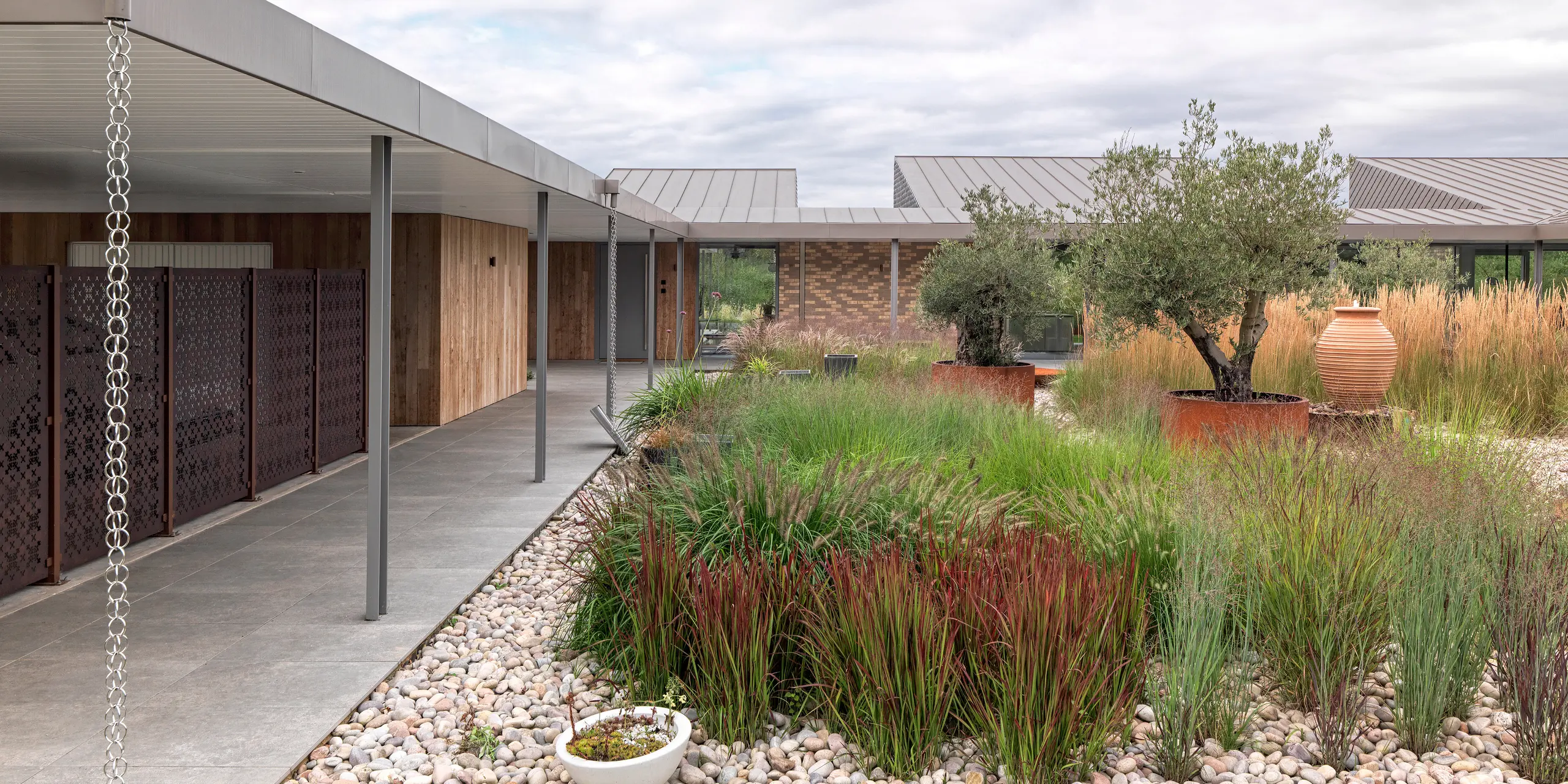 Vue détaillée d'une maison moderne du West Sussex avec toit plat en aluminium PREFALZ couleur  P.10bronze. On y découvre une terrasse couverte et clairement structurée, avec des colonnes fines, des chaînes d'évacuation des eaux pluviales intégrées avec style et un jardin varié composé de graminées ornementales, d'oliviers et de pierres naturelles. Les toitures durables en aluminium PREFA soulignent les lignes modernes et s'intègrent harmonieusement au concept architectural durable.