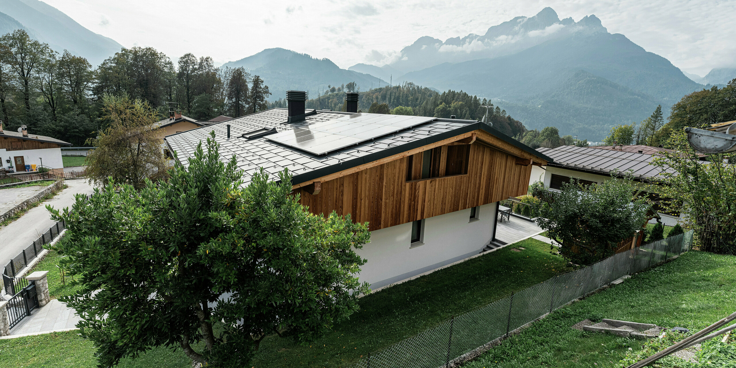 Rückseite eines sanierten Einfamilienhauses in Agordo, Italien. Das neue Dach wurde mit PREFA Dachplatten R.16 in P.10 Anthrazit eingedeckt. Das widerstandsfähige Aluminiumdach bietet optimalen Schutz vor extremen Witterungseinflüssen und ist mit einer nachhaltigen PV-Anlage ausgestattet. Die PREFA Dachentwässerung mit Kastenrinne und Ablaufrohr sorgt für eine zuverlässige Wasserableitung. Die Kombination aus naturbelassener Holzfassade, weißer Putzfassade und langlebigem Aluminiumdach fügt sich harmonisch in die alpine Landschaft ein.