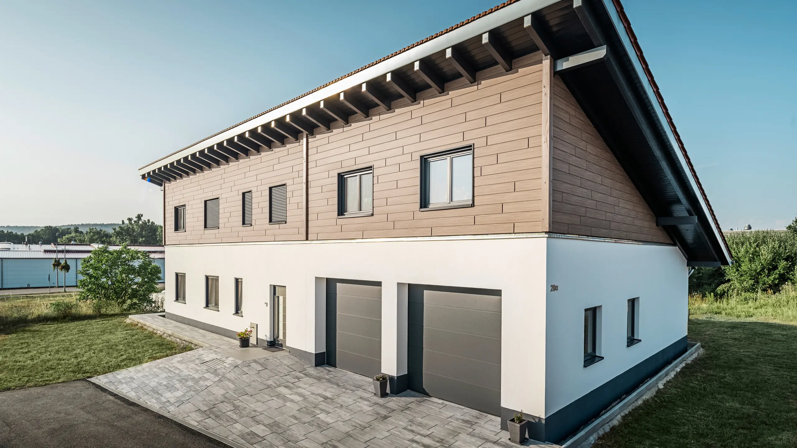 Vue latérale d'une maison individuelle avec un toit à une pente à Bruck. L'étage supérieur est enveloppé de Sidings PREFA en noyer foncé. Sur le côté droit de l'image, on peut voir deux portes de garage en anthracite. Au centre du bâtiment se trouve la porte d'entrée. Le socle est d'un blanc sobre. 