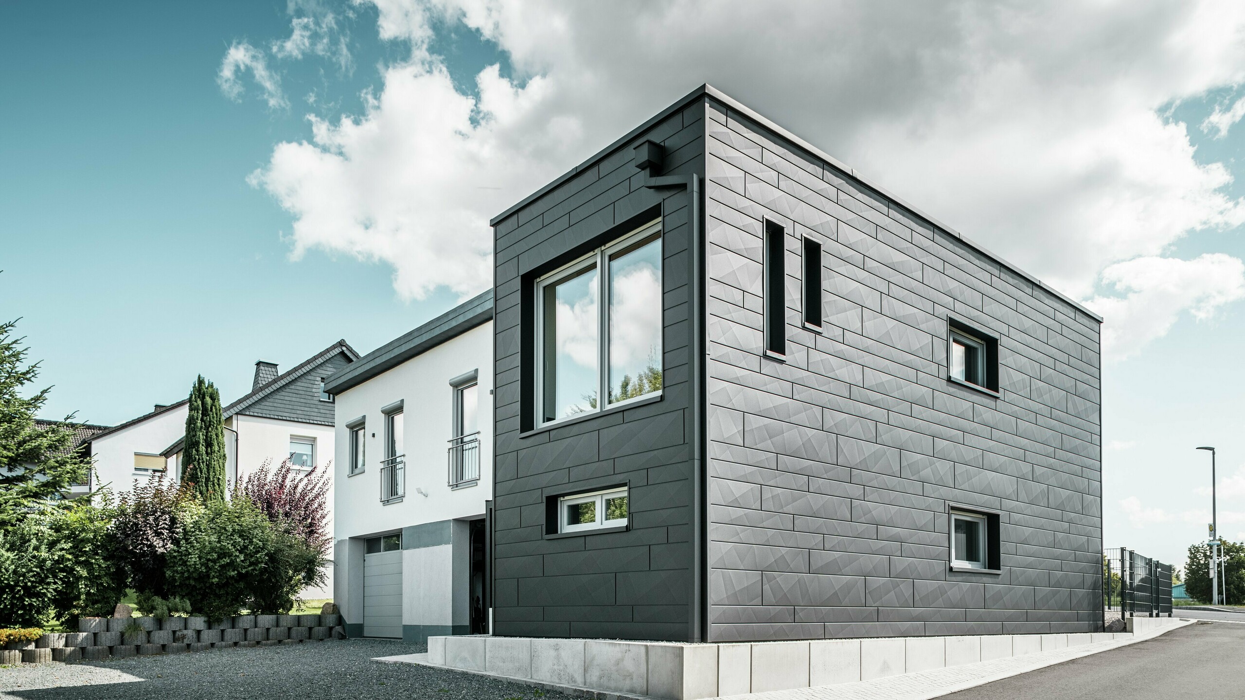 Die stilvolle und moderne Fassade des sanierten Einfamilienhauses in Teuschnitz zeigt die klar strukturierten PREFA Siding.X Paneele in P.10 Schwarz. Die geometrische Anordnung der hochwertigen Blechpaneele verleiht dem Gebäude eine markante, zeitlose Ästhetik. Die Fenster sind von schwarzen Aluminium Verbundplatten umrahmt, was das Design harmonisch abrundet. Die integrierte PREFA Dachentwässerung mit dem Quadratrohr sorgt für eine funktionale und ästhetische Lösung.