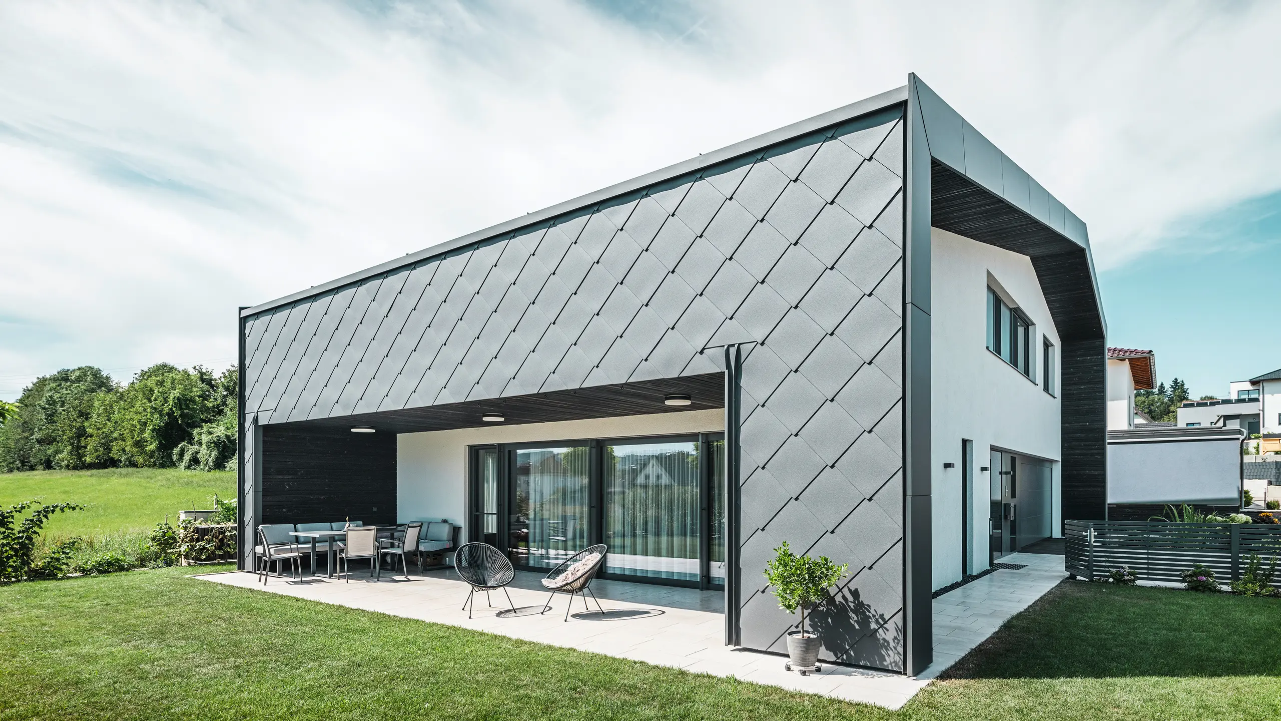 Jardin bien entretenu d'une maison unifamiliale à Aschach. Le bâtiment est élégamment recouvert de losanges de façade 44x44 gris sombre. La terrasse couverte offre un coin salon confortable avec vue sur la pelouse verte luxuriante. Le revêtement élégant en aluminium associé à des surfaces en plâtre blanc garantit une architecture moderne et résistante aux intempéries. De grandes fenêtres laissent entrer beaucoup de lumière du jour à l'intérieur et créent une atmosphère ouverte et lumineuse.