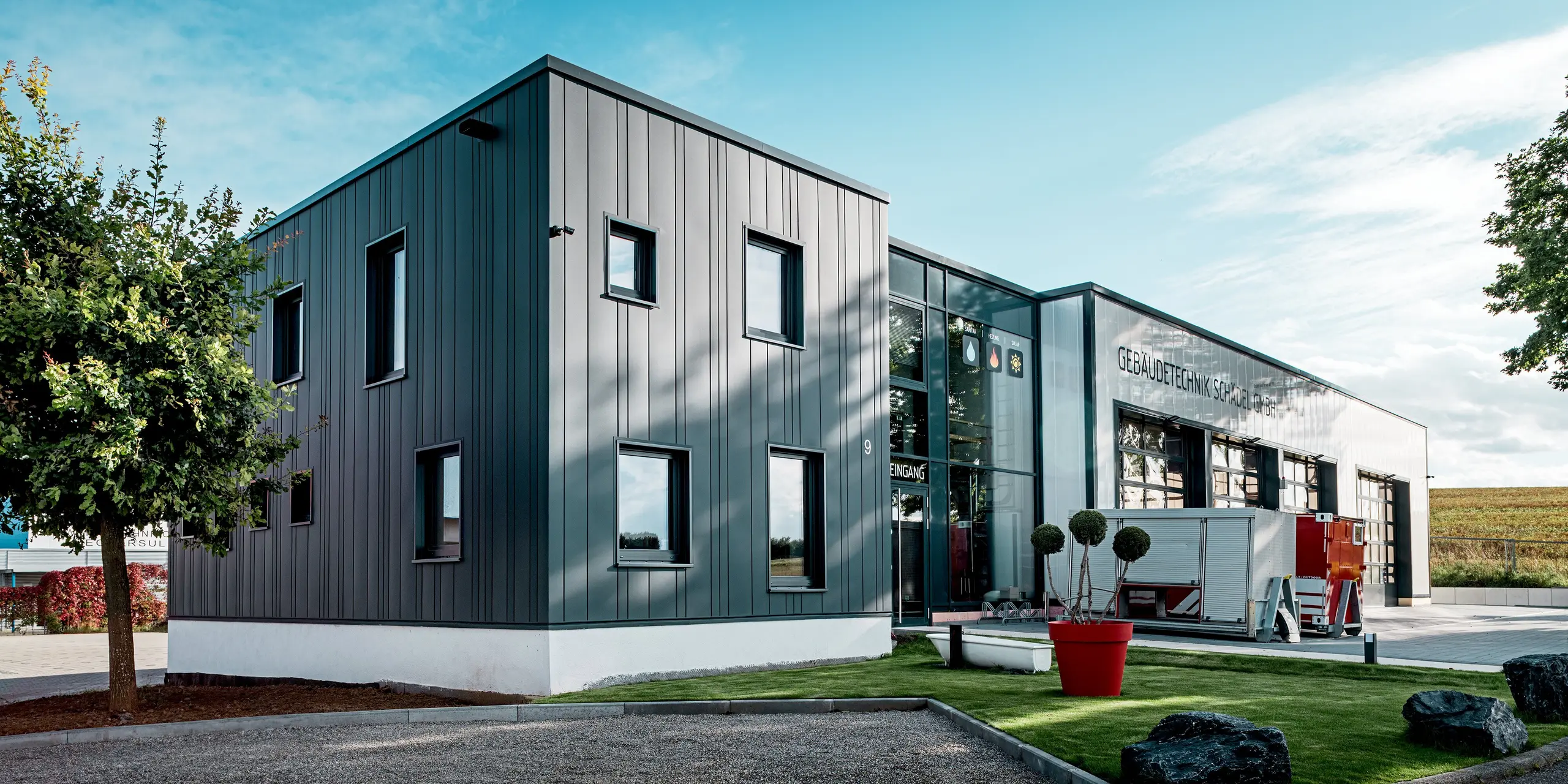 La scène met en valeur le bâtiment aux lignes épurées de Schädel GmbH, entreprise spécialisée dans les technologies du bâtiment, avec sa façade remarquable en tôle PREFA, dont les lignes verticales anthracite attirent immédiatement le regard. Le jeu de largeurs de Sidings variées confère au bâtiment un rythme visuel. L'alliance de l'aluminium, de la façade vitrée et du parvis ouvert crée un effet moderne et saisissant. La surface PREFA, d'une grande durabilité, résiste parfaitement aux intempéries et à l'usure.