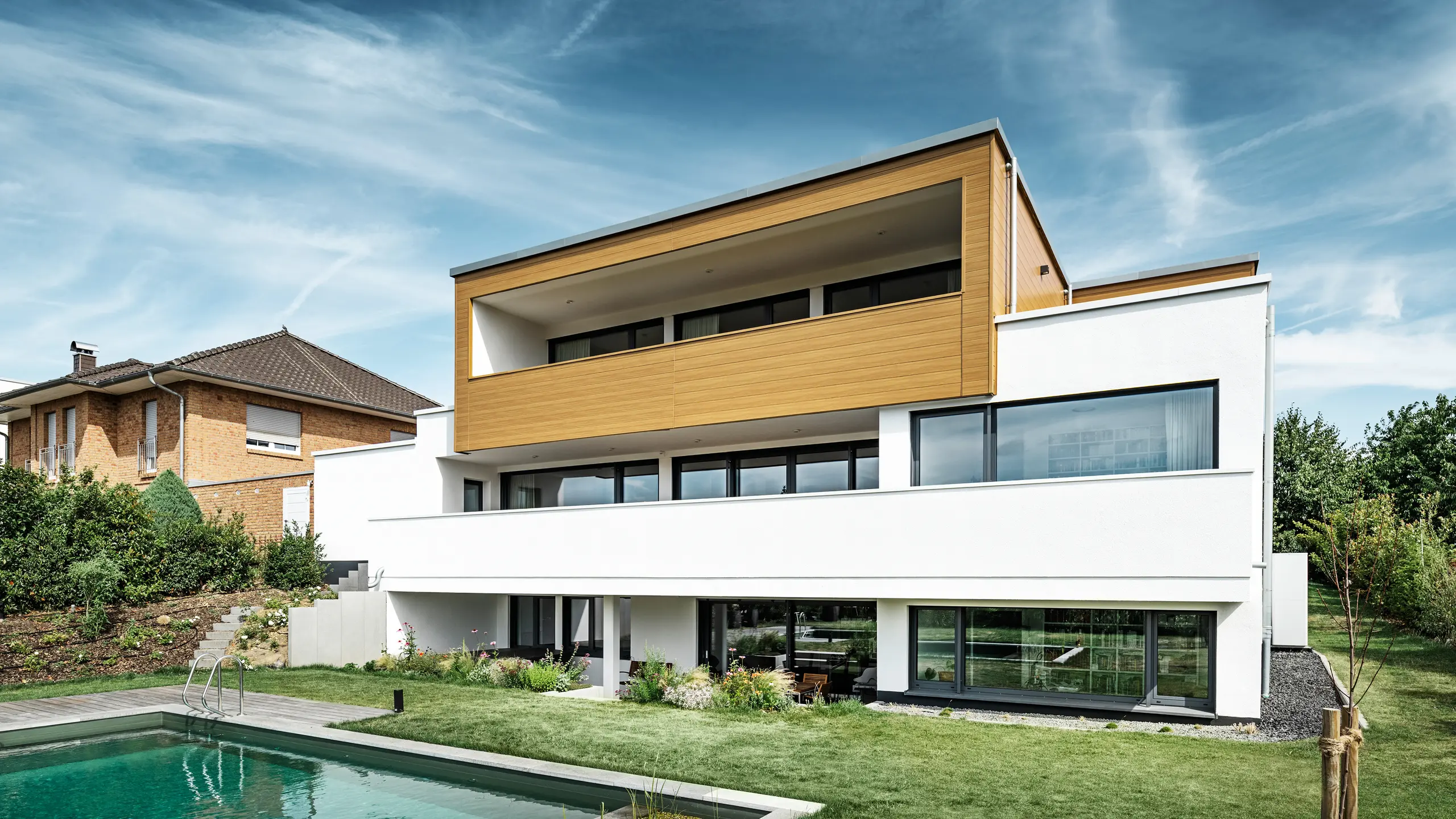 Die Rückseite eines modernen Einfamilienhauses in Usingen, Deutschland. Am unteren Bildrand ist die grüne Wiese, ein Pool sowie ein junger Gartenbaum zu sehen. Das auffällige Haus verfügt über einen weißen Gebäudesockel aus einer Putzfassade, einer Aluminiumfassade am Obergeschoss aus PREFA Sidings in Eiche natur sowie großen Fensterflächen mit einem anthrazitfarbenen Rahmen. Aus der Distanz ist das Nachbarhaus mit einer Ziegelfassade am linken Bildrand zu erkennen.