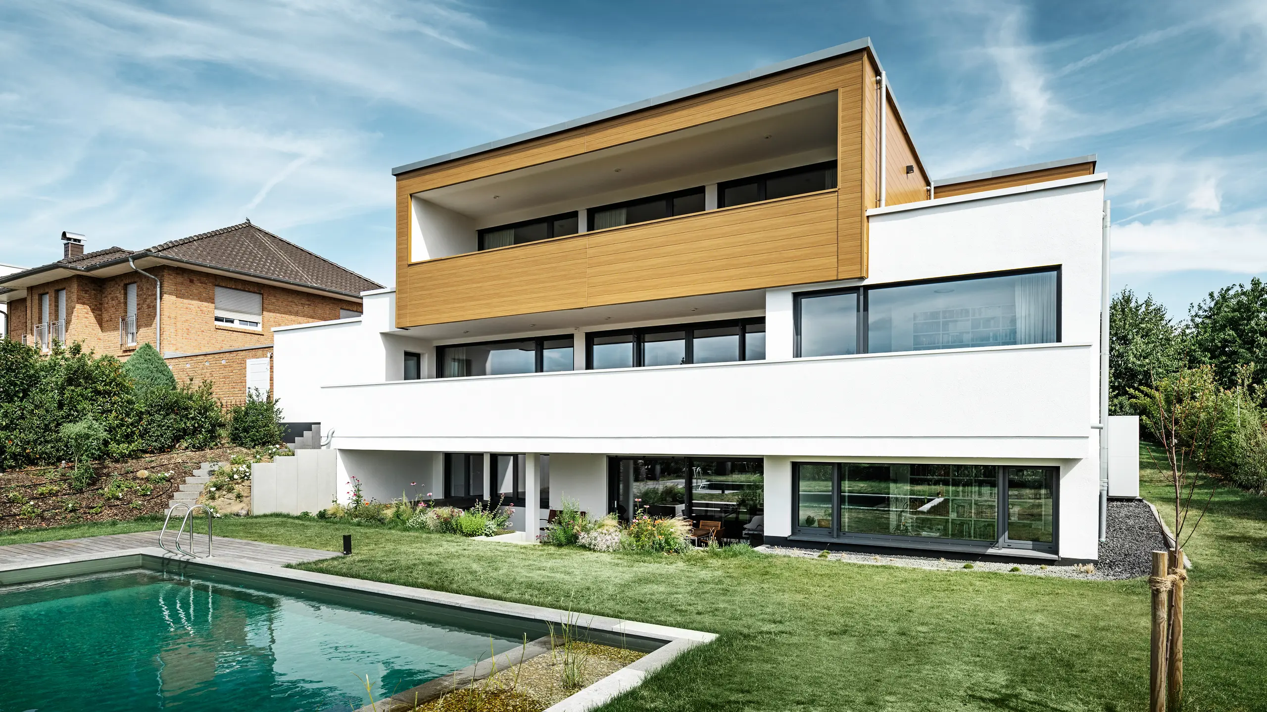 L'arrière d'une maison individuelle moderne à Usingen, Allemagne. En bas de l'image, on peut voir une pelouse verte, une piscine ainsi qu'un jeune arbre de jardin. Cette maison remarquable dispose d'un socle de bâtiment blanc composé d'une façade en crépi, d'une façade en aluminium à l'étage supérieur en PREFA Sidings en chêne naturel ainsi que de grandes surfaces de fenêtres avec un cadre de couleur anthracite. De loin, on distingue la maison voisine avec une façade en briques sur le bord gauche de l'image.