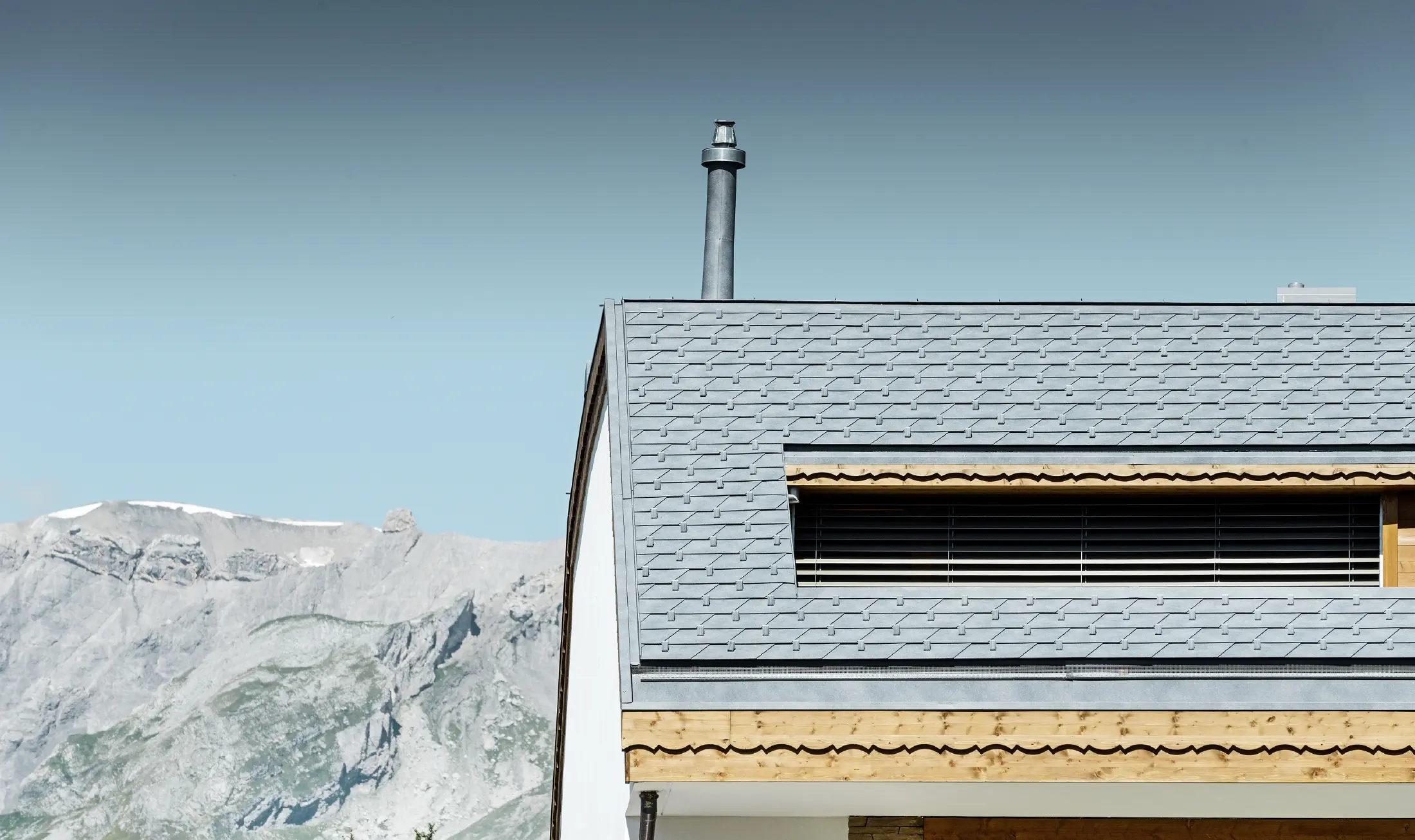 Immeuble d’habitation de Crans-Montana (Suisse) avec les Alpes en arrière-plan — Façade intégrant de nombreux éléments en bois et dont la toiture a été réalisée avec des bardeaux en aluminium PREFA de couleur gris pierre