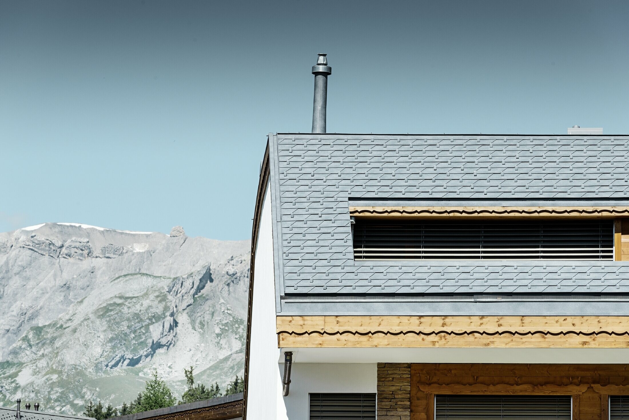 Immeuble d’habitation de Crans-Montana (Suisse) avec les Alpes en arrière-plan — Façade intégrant de nombreux éléments en bois et dont la toiture a été réalisée avec des bardeaux en aluminium PREFA de couleur gris pierre