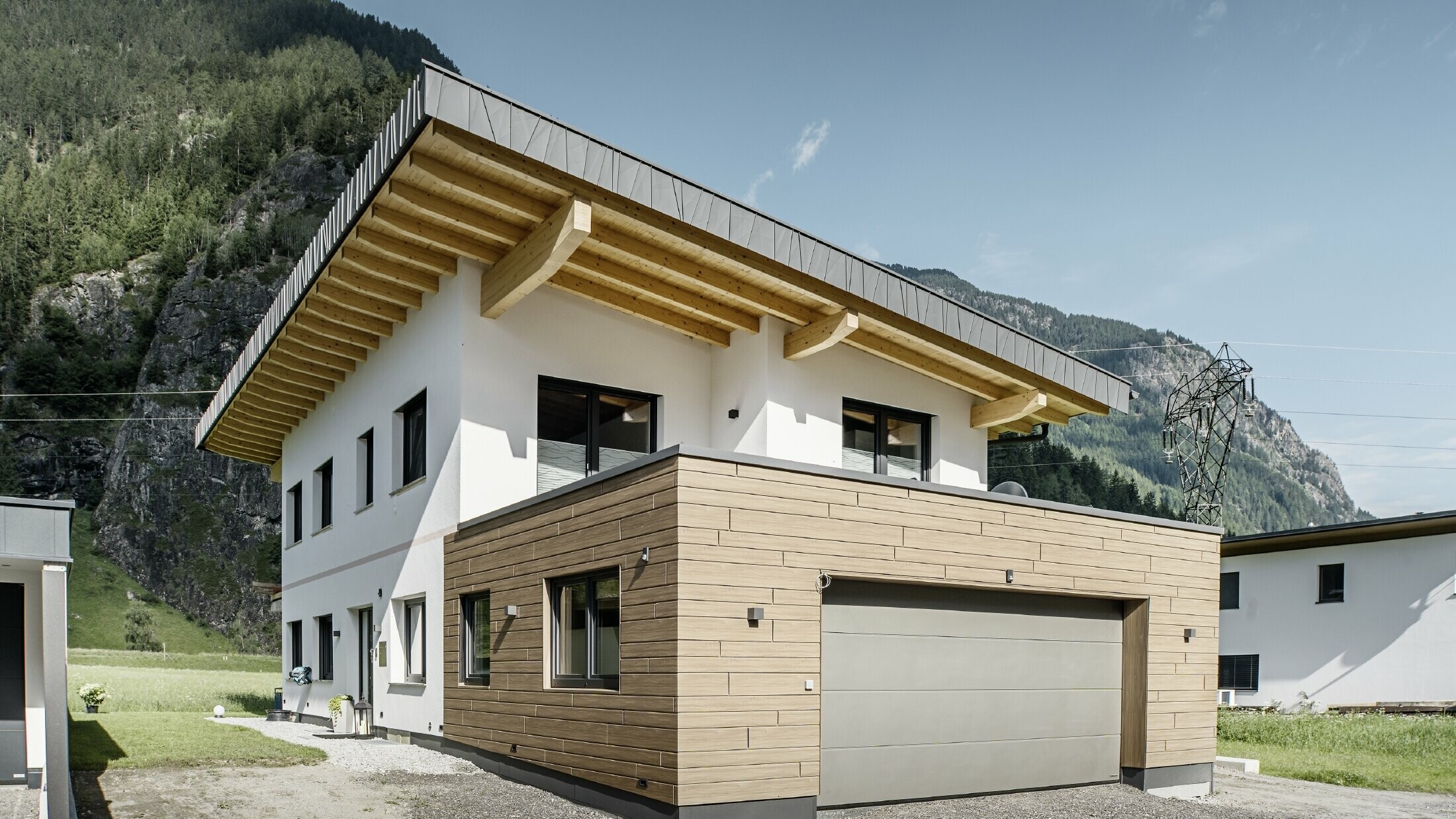 Maison individuelle moderne avec rénovation de façade : façade extérieure rénovée avec un revêtement horizontal aspect bois, combiné à des surfaces crépies en blanc, un revêtement de garage intégré et une architecture contemporaine dans un environnement alpin.