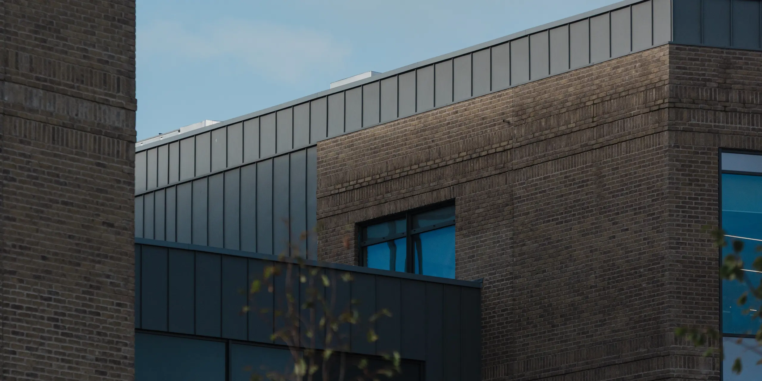 Une vue précise de la façade du Northern Regional College de Ballymena révèle que la partie supérieure du bâtiment est coiffée de panneaux PREFALZ en aluminium à joints debout, gris patiné, installés verticalement. La pureté des lignes de la bande d'aluminium contraste harmonieusement avec la chaleur de la brique et les ouvertures de fenêtres profondément encastrées. La surface métallique, d'une grande sobriété, reflète subtilement la lumière du jour sur le ciel bleu. L'aluminium PREFA impressionne ici par sa durabilité, sa précision formelle et son caractère intemporel, offrant une solution nécessitant peu d'entretien pour les bâtiments scolaires modernes.