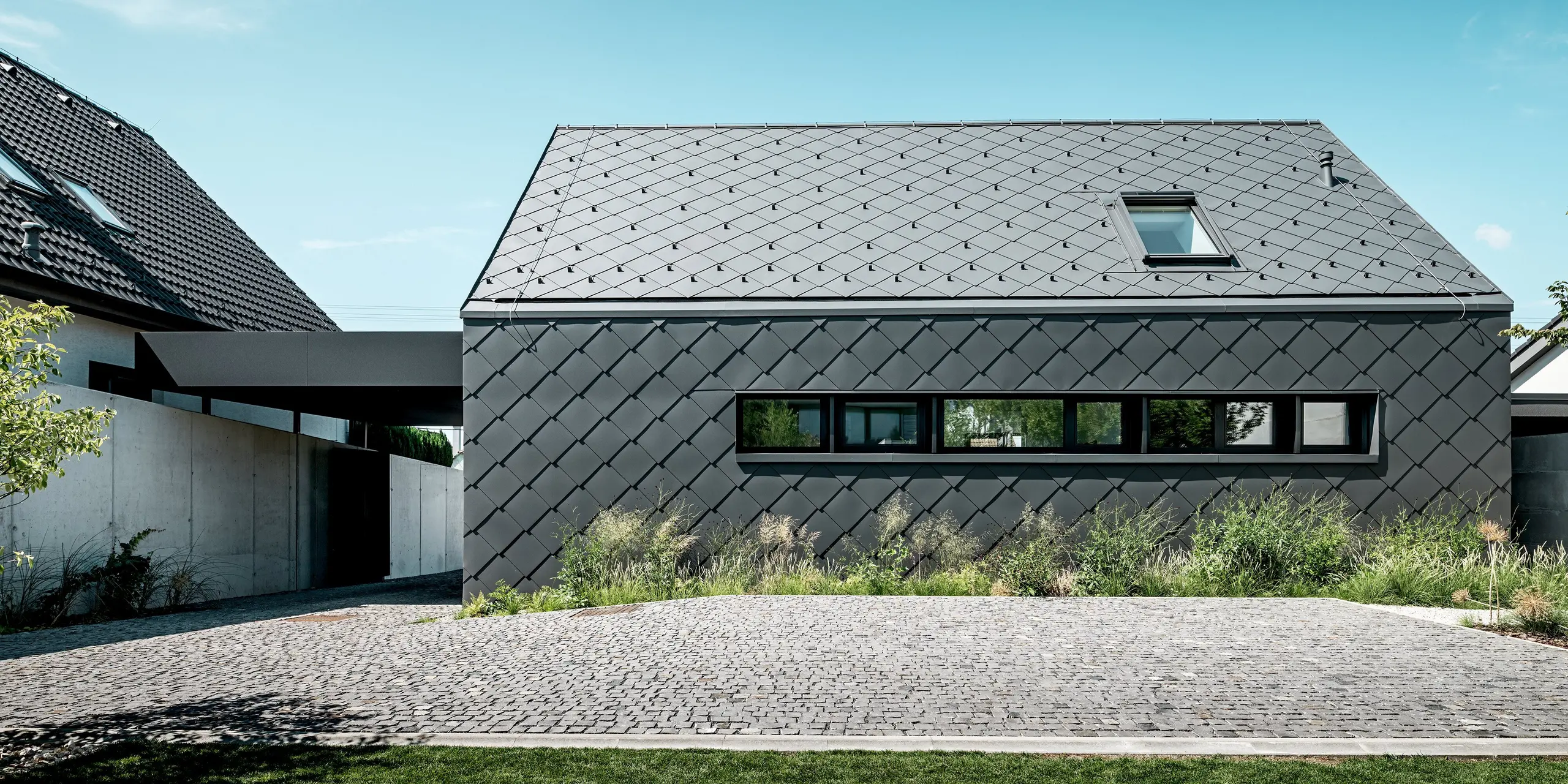 L'image présente la vue latérale d'une maison individuelle moderne à Trnava-Modranka, entièrement revêtue de losanges de toiture et de faça de 44x44 PREFA, coloris P.10 gris sombre. Le motif régulier des losanges confère à la toiture et à la façade une surface sobre et élégante, et souligne la précision du design. Ces produits en aluminium durable offrent une excellente protection contre les intempéries, une couleur inaltérable et un aspect robuste et moderne. La végétation naturelle à l'avant crée un contraste doux et agréable avec le revêtement métallique.