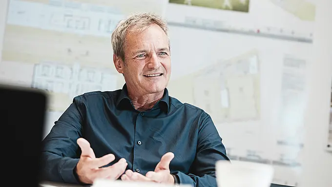 Portrait de l'architecte Max Stoisser, associé fondateur de planconsort, dans une conversation. Il porte une chemise bleu foncé et fait des gestes pendant qu'il parle. En arrière-plan, vous pouvez voir des plans et des visualisations du centre événementiel ZIB Nikolai.