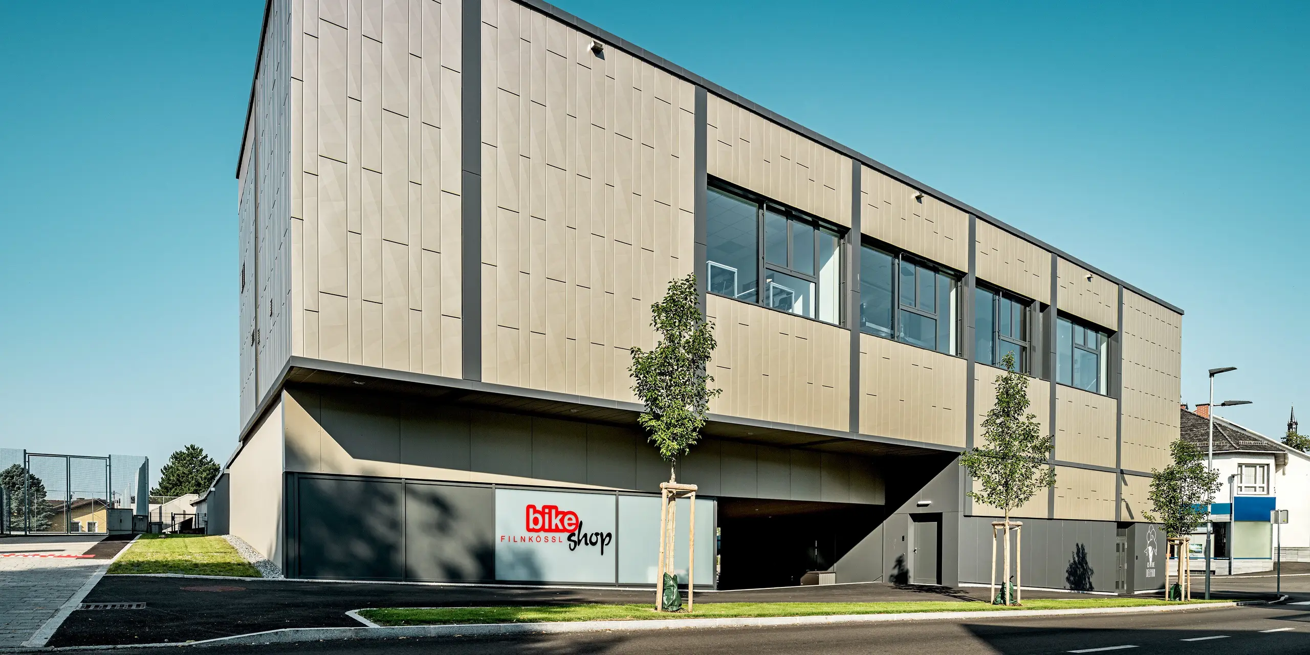 L'image présente le centre commercial moderne de Rohrbach, reconnaissable à sa façade en aluminium composée de Siding.X PREFA couleur bronze. Le complexe impressionne par son design contemporain. Les éléments distinctifs de la façade créent non seulement un effet visuel remarquable, mais offrent également une protection efficace contre les intempéries. Le magasin de vélos Filnkössl et le glacier Delphin proposent diverses activités de loisirs dans cette petite ville de Basse-Autriche.
