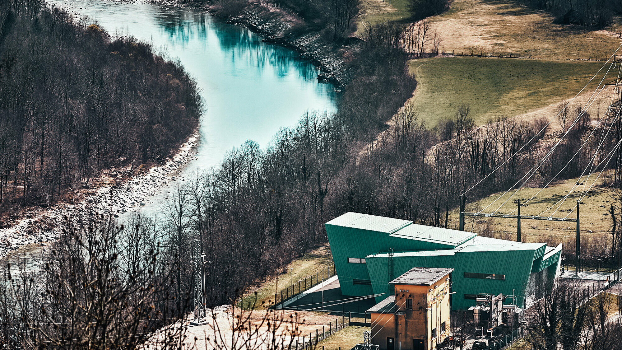 Das Umspannwerk aus der Vogelperspektive samt Umgebung, die gegenläufigen Dachflächen kommen zur Geltung, das alte Umspannwerk steht dicht daneben. Die Farbe der Fassade deckt sich mit jener der vorbeifließenden Soča.