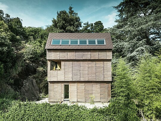 Architecture moderne en France - maison avec toiture en bardeaux PREFA de couleur P.10 brun noisette et façade en bois de saule, située sur une colline escarpée avec puits de lumière spacieux.