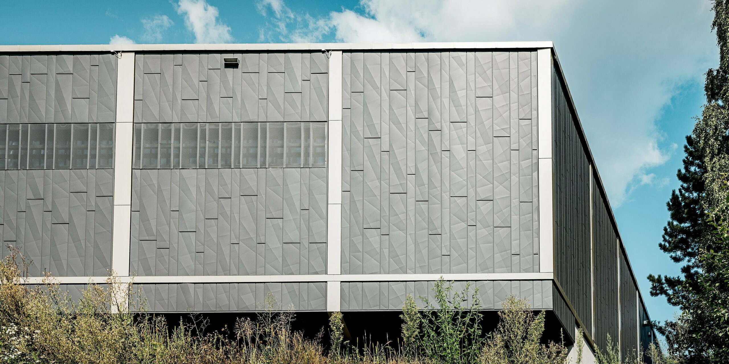 Das Bild zeigt die Rückseite des Einkaufszentrums in Rohrbach mit einer modernen Blechfassade aus PREFA Siding.X-Paneelen in Anthrazit. Die geometrischen Strukturen der Paneele schaffen eine dynamische Textur, die dem Gebäude eine markante und zeitgenössische Ästhetik verleiht. Besonders auffällig sind die horizontalen Streifen aus perforierten Elementen, die nicht nur eine optische Vielfalt bieten, sondern auch für eine gezielte Belüftung und Lichtdurchlässigkeit sorgen. Die Fassade fügt sich harmonisch in die umgebende Natur ein, wobei das Spiel zwischen den satten grauen Tönen und dem grünen Bewuchs eine reizvolle Verbindung zwischen Architektur und Landschaft bildet.