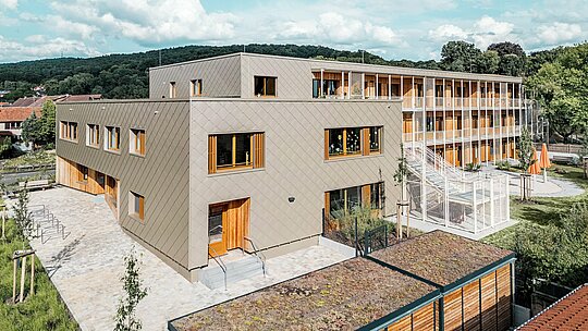 Vue d'en haut, l'image révèle le nouveau bâtiment en forme de L de la crèche d'Alzenau, avec son aile résidentielle attenante : un bâtiment hybride et novateur, construit selon des méthodes durables. La façade en aluminium PREFA, couleur bronze, composée de losange de façade 44 x 44, définit l'extérieur moderne et structuré de la crèche. De larges fenêtres en bois et des persiennes verticales créent une atmosphère accueillante et chaleureuse. L'espace arrière, avec son escalier extérieur, ses aires de jeux et ses espaces verts, illustre la séparation fonctionnelle et la connexion simultanée des différents usages. Le toit végétalisé et l'association de matériaux – aluminium, bois et béton – témoignent des exigences écologiques élevées du projet et affirment une vision forte de l'architecture éducative et résidentielle durable.