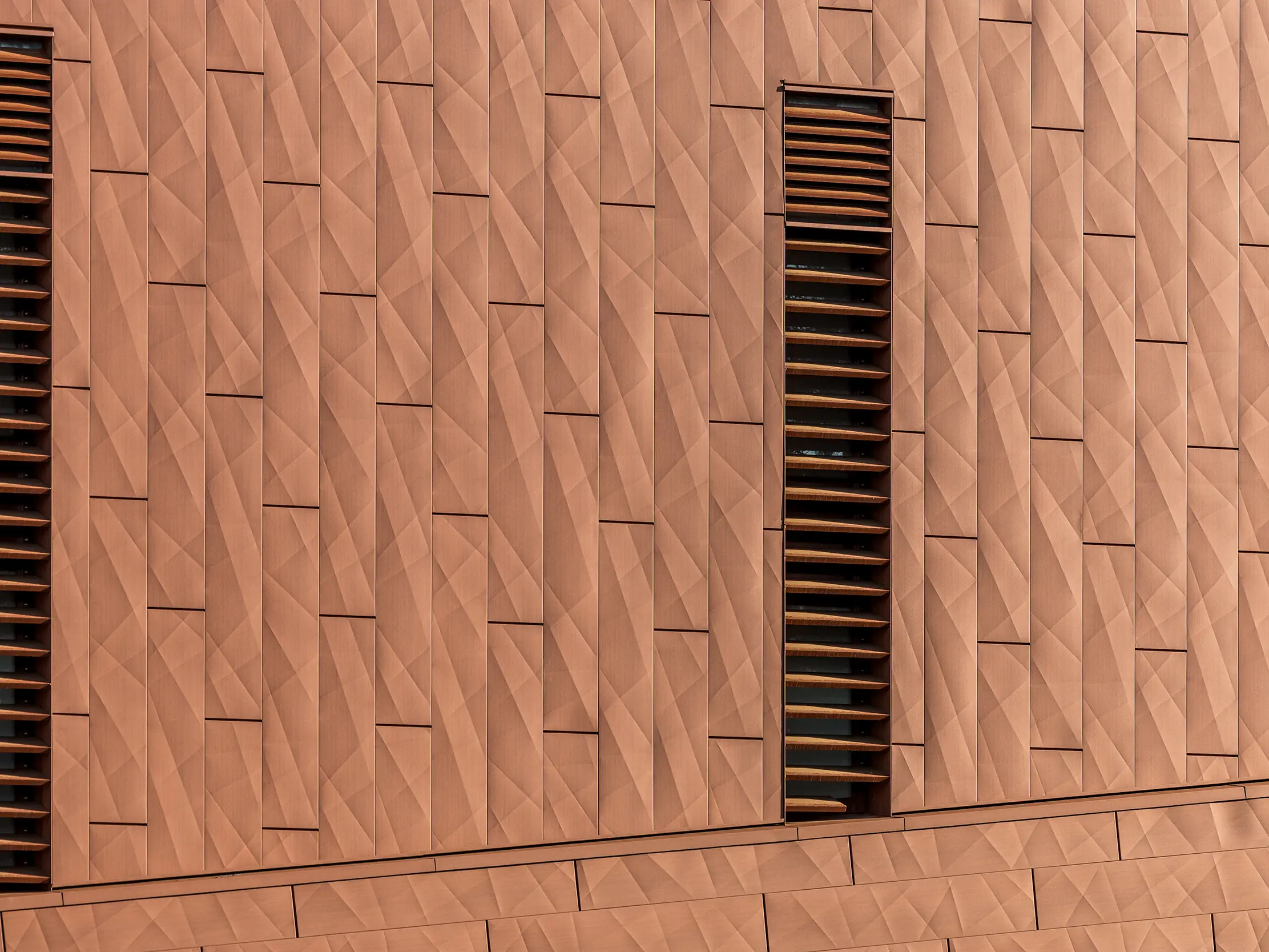 Détail de la façade en aluminium PREFA Siding.X de la station de pompage d'eau potable WML à Lottum. Sa surface géométrique finement plissée présente un aspect chaleureux, évoquant la rouille. L'agencement précis des panneaux crée un jeu d'ombre et de lumière saisissant, tandis que les fines ouvertures verticales à persiennes structurent la façade et constituent des éléments visuels marquants. Légers, durables et entièrement recyclables, les panneaux en aluminium PREFA offrent une résistance exceptionnelle aux intempéries, une excellente tenue des couleurs et une enveloppe de bâtiment durable et nécessitant peu d'entretien.