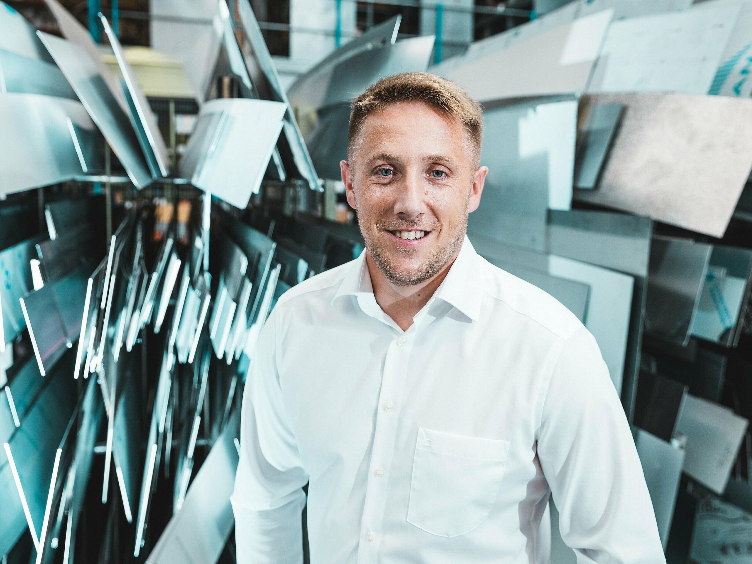 Confiant et affable, Marcel Schmidt, chef de projet chez ASA Schüßler, entreprise spécialisée dans les toitures métalliques, se tient devant un impressionnant mur composé d'éléments en aluminium de formes variées. Les panneaux métalliques, agencés avec dynamisme, reflètent efficacement la lumière et symbolisent l'innovation, la précision et la qualité du travail des métaux. Cet homme en chemise blanche incarne la compétence et l'excellence de la production moderne. À l'arrière-plan, on découvre la diversité des possibilités de transformation de l'aluminium : des systèmes de toiture et de façade aux solutions spéciales sur mesure.