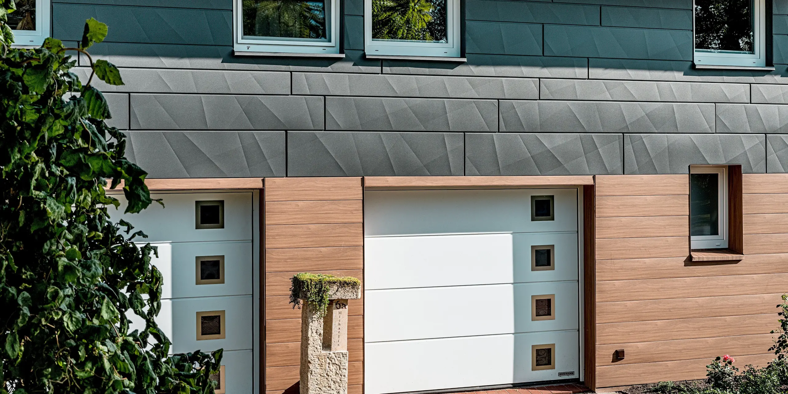 Vue détaillée de la façade rénovée d'une maison individuelle revêtue d'un bardage en aluminium PREFA de haute qualité. L'attention se porte sur les portes de garage blanches, encadrées par un bardage PREFA couleur noyer foncé à l'aspect bois authentique, qui confère chaleur et profondeur à l'entrée. Au-dessus, le bardage PREFA Siding.X P.10 gris sombre, aux arêtes longitudinales et transversales marquées, apporte une touche de modernité et crée un jeu d'ombres et de lumières saisissant. La pureté des lignes horizontales, associée à la durabilité, à la résistance aux intempéries et à la facilité d'entretien des panneaux d'aluminium, fait de cette rénovation de façade un exemple remarquable d'architecture contemporaine et de qualité durable.