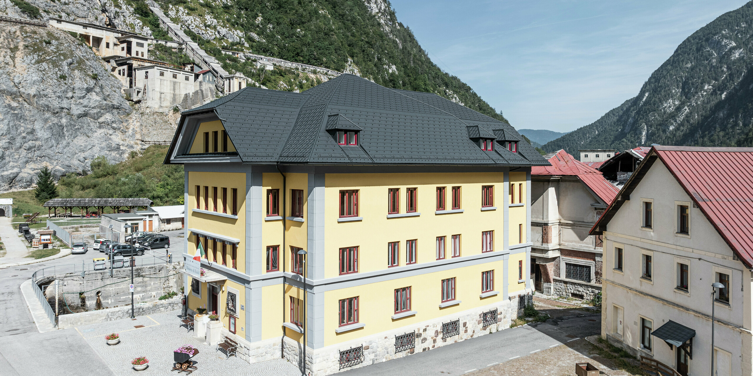 Historisches Museumsgebäude Museo Cave del Predil in Italien mit gelber Fassade und anthrazitfarbenem PREFA Aluminiumdach (Dachplatte P.10 anthrazit). Im Hintergrund beeindruckende Bergkulisse und alpine Umgebung. Das Dach überzeugt durch Langlebigkeit und Robustheit - ideal für extreme alpine Witterungsbedingungen.