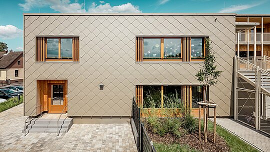 La façade arrière de la crèche moderne d'Alzenau, vue de face, impressionne par ses lignes épurées et son élégante façade composée de losanges de façade 44x44 PREFA couleur bronze. La structure géométrique de la façade crée un motif harmonieux, évoquant des écailles, accentué par l'horizontalité apaisante des fenêtres. De généreux encadrements en bois et des persiennes verticales apportent une touche naturelle et créent une atmosphère chaleureuse et accueillante. L'alliance judicieuse de l'aluminium durable et du bois local témoigne d'une architecture durable et d'une grande qualité de conception. L'image met en lumière la précision de l'exécution, le choix judicieux des matériaux et l'intégration réussie du bâtiment dans son environnement urbain.