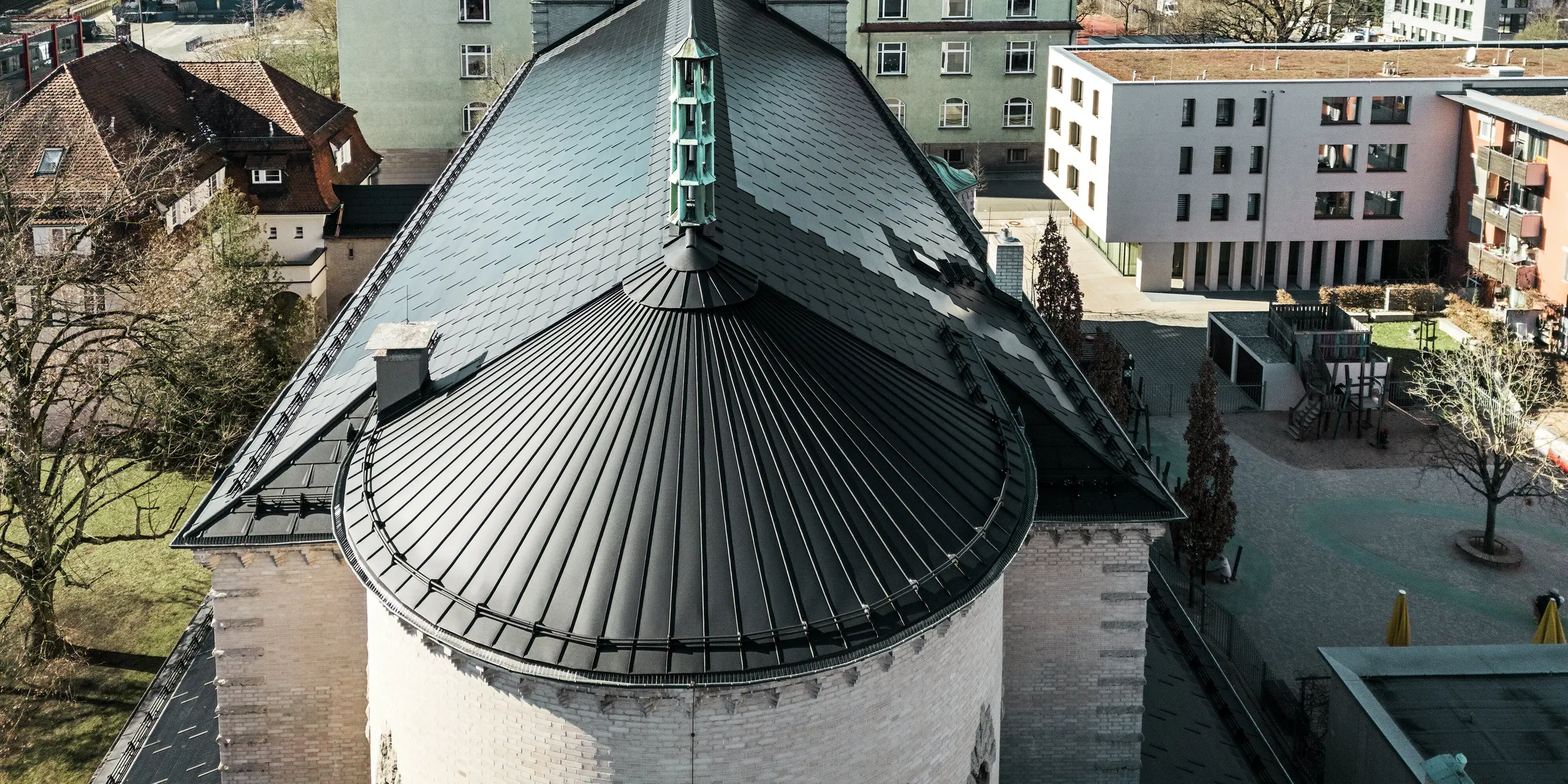 L'image montre le toit arrondi et saisissant à l'arrière de l'église Saint-Charles-Borromée de Nuremberg. La courbe du bâtiment a été recouverte d'une toiture à joint debout en aluminium noir. Le toit à deux versants, qui protège la partie principale du bâtiment, a été recouvert de tuiles solaires PREFA noires. Ces tuiles solaires innovantes constituent un point fort visuel et produisent simultanément de l'électricité à partir d'énergies renouvelables. La vue sur le toit et la flèche centrale illustre l'intégration harmonieuse de la technologie solaire moderne dans l'architecture historique. À l'arrière-plan, on distingue des bâtiments traditionnels et modernes, soulignant la combinaison réussie entre préservation du patrimoine et construction durable.