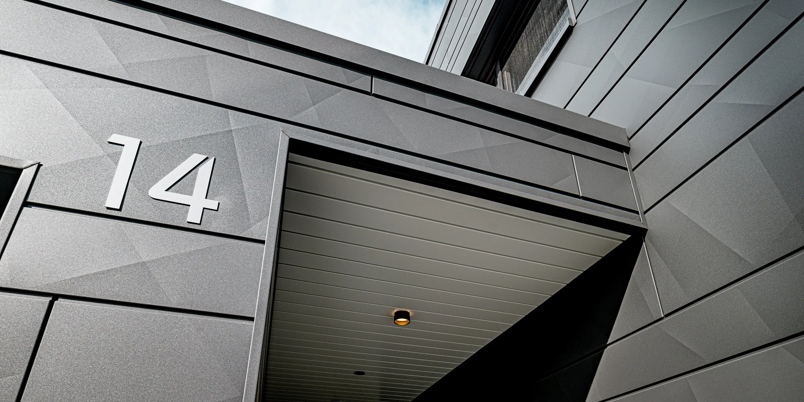Vue détaillée de l'entrée couverte d'une maison individuelle moderne à Heidenheim, revêtue d'une façade Siding.X PREFA couleur P.10 gris sombre. Les panneaux d'aluminium, disposés selon différents angles, créent un motif géométrique saisissant, jouant avec les ombres et les reflets. Le numéro « 14 » se détache nettement sur la surface robuste. L'auvent protecteur, équipé de spots encastrés, souligne la qualité de la réalisation et démontre la durabilité et la résistance aux intempéries de la solution aluminium PREFA.